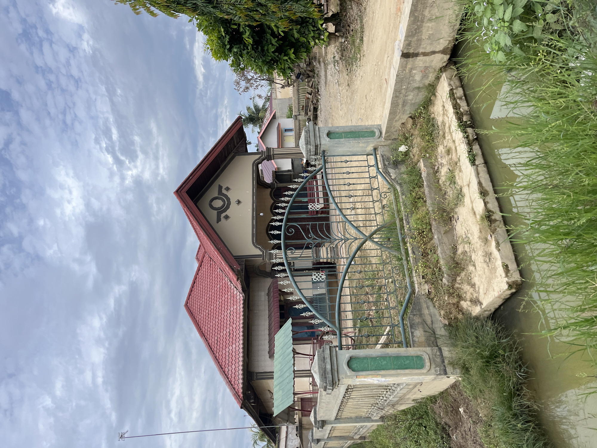 The image depicts a residential property in Bundar, Indonesia. The foreground features a narrow, muddy canal with overgrown green grass along its banks. A concrete pathway leads from the canal towards a house, with steps made of concrete. A decorative metal gate, painted green, is a prominent feature of the property's fence, which is constructed from concrete pillars and horizontal bars, also painted green. The house itself has a prominent red tiled roof and a beige-colored facade. A smaller green awning is visible on the left side of the house, and a larger entrance with decorative columns is on the right. The sky is overcast with clouds. In the background, other houses and trees are visible, suggesting a semi-rural or suburban setting.