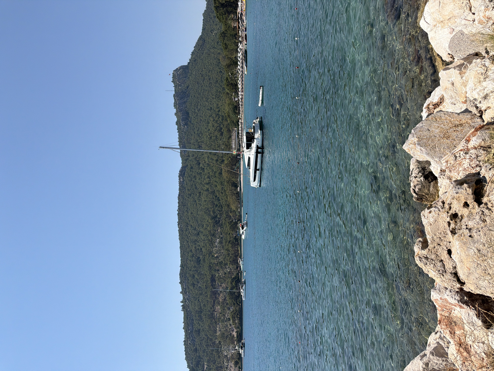 This is a landscape-oriented photograph of a coastal scene with a clear, turquoise body of water in the foreground and a lush, green, tree-covered mountain in the background. The water is calm, with visible ripples reflecting the sunlight. On the right side of the image, a rocky shoreline with large, rough-textured stones is prominent.
Several boats are moored in the water, including a white catamaran with a tall mast in the center of the frame. There are also smaller boats and buoys visible further out. In the distance, along the base of the mountain, a line of white buildings and a beach with umbrellas can be seen, suggesting a populated area or resort. The sky is a clear, bright blue, indicating fair weather and likely daytime. The overall impression is one of a peaceful and scenic coastal environment. The image appears to have been taken from the shore, looking out towards the water and the mountain.
