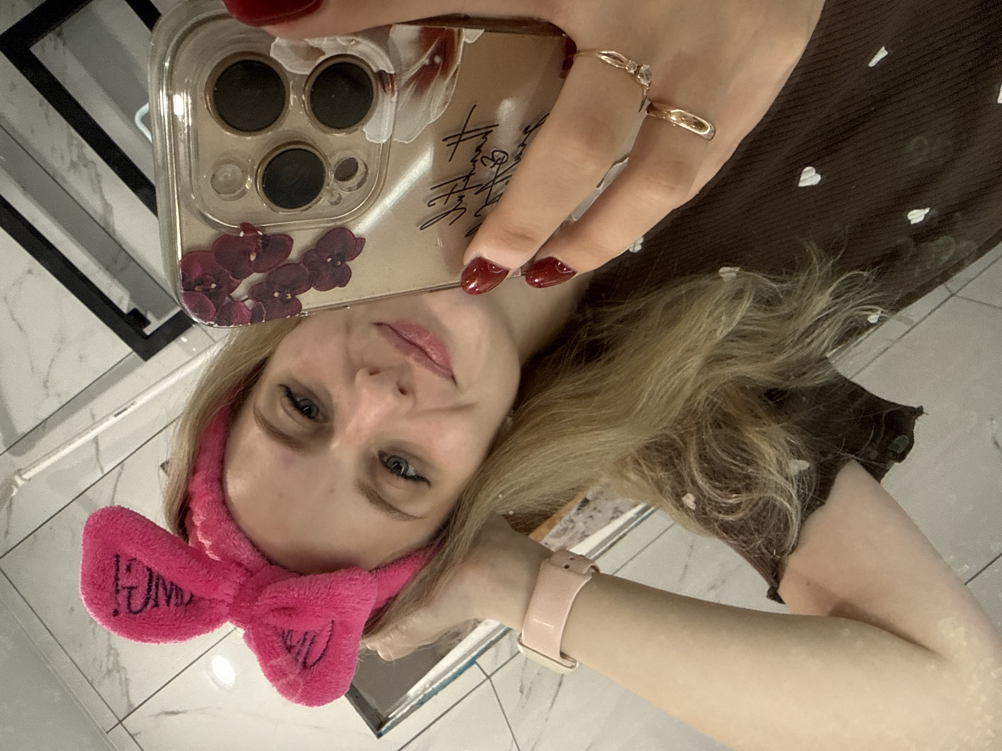 A young woman with blonde hair and blue eyes is taking a selfie in a bathroom. She is wearing a pink fuzzy headband with a bow and text, a brown top with a heart pattern, and a light pink smartwatch. She has on red nail polish and two rings on her right hand. Her phone, with a clear case adorned with floral patterns, is visible in her hand as she takes the picture. The bathroom has tiled walls and a mirror reflecting the scene.