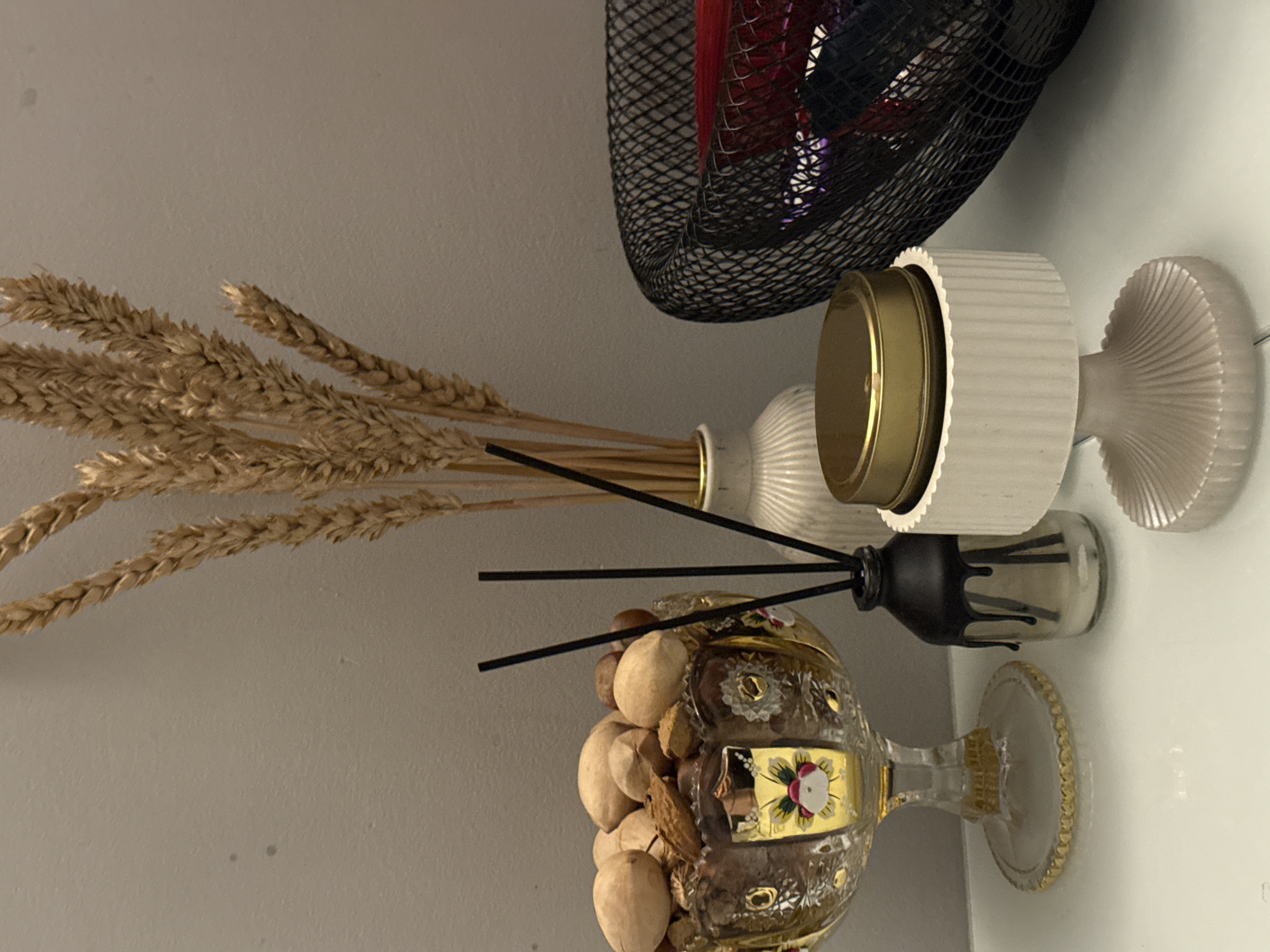 The image shows a still life composition with various decorative objects arranged on a white surface against a muted gray wall.
Objects include:
* A bouquet of dried wheat stalks in a white ceramic vase.
* A decorative glass goblet filled with nuts.
* A black diffuser with several black reeds.
* A white, ribbed candle holder with a golden lid.
* A black mesh basket-like object containing colorful objects (likely fabrics or other decorative items).
The arrangement appears to be a styled display, possibly in a residential setting. There are no people visible, and the scene is static. The environment is a neutral indoor space, with the focus solely on the arrangement of the objects. The city and state are unknown.