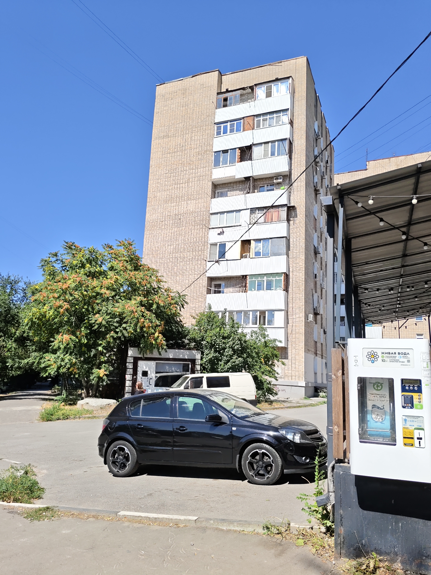 A black hatchback car is parked on a paved surface next to a building with a light brown brick facade and white balconies. A large green tree with clusters of orange-red fruits is to the left of the car. In the foreground, there is a white vending machine with Russian text, and a portion of a covered walkway with string lights is visible to the right. The sky is clear and blue. The scene appears to be in an urban environment, possibly a residential area in Rostov-on-Don, Russia.