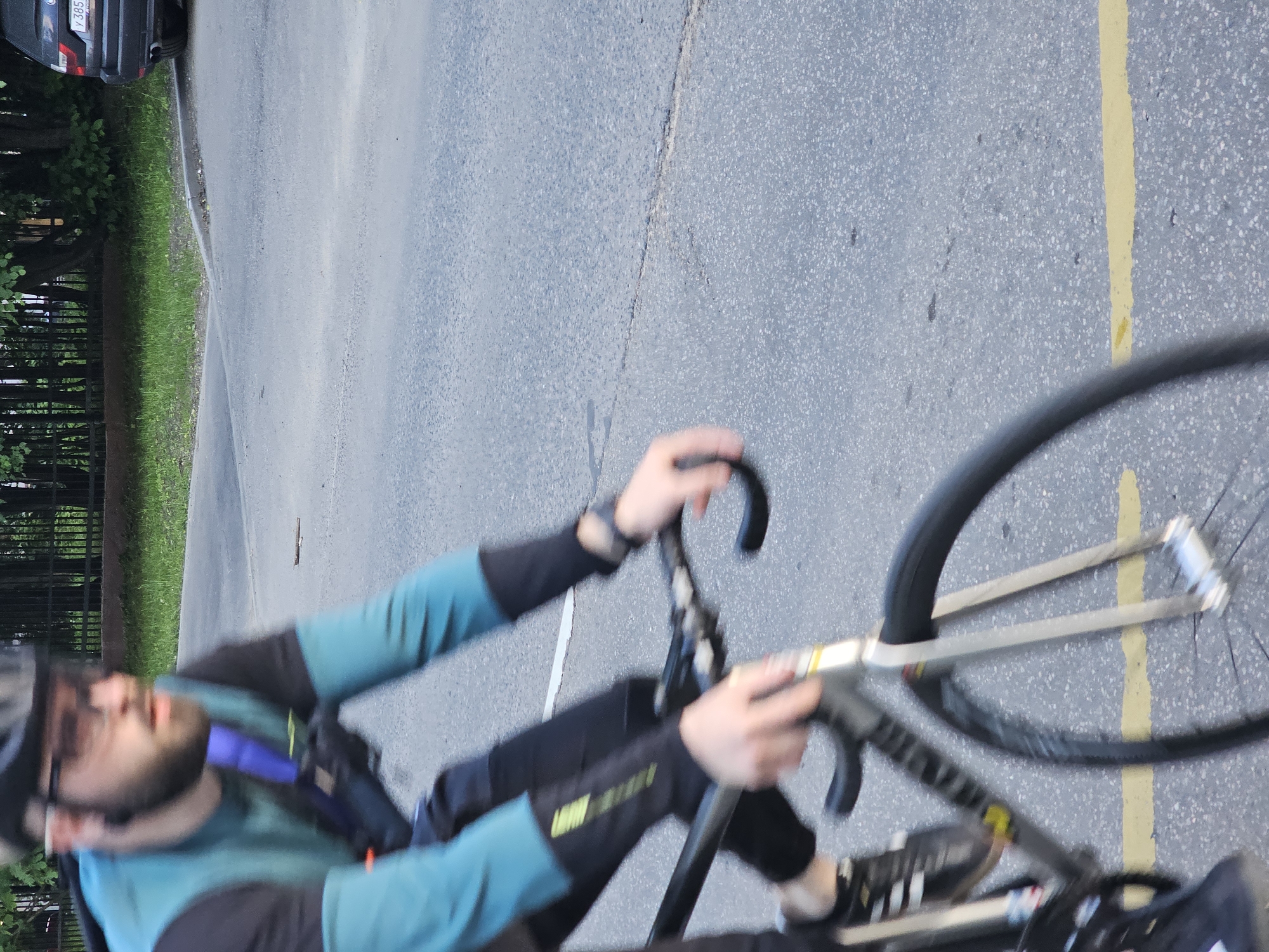A cyclist is riding on an asphalt road marked with a yellow line. The rider is wearing a helmet, glasses, and cycling apparel with black and turquoise accents. A blurred background includes vegetation and a car partially visible at the edge of the road. The location is likely Khimki, Russia, based on the license plate of the car, although this is not explicitly depicted.