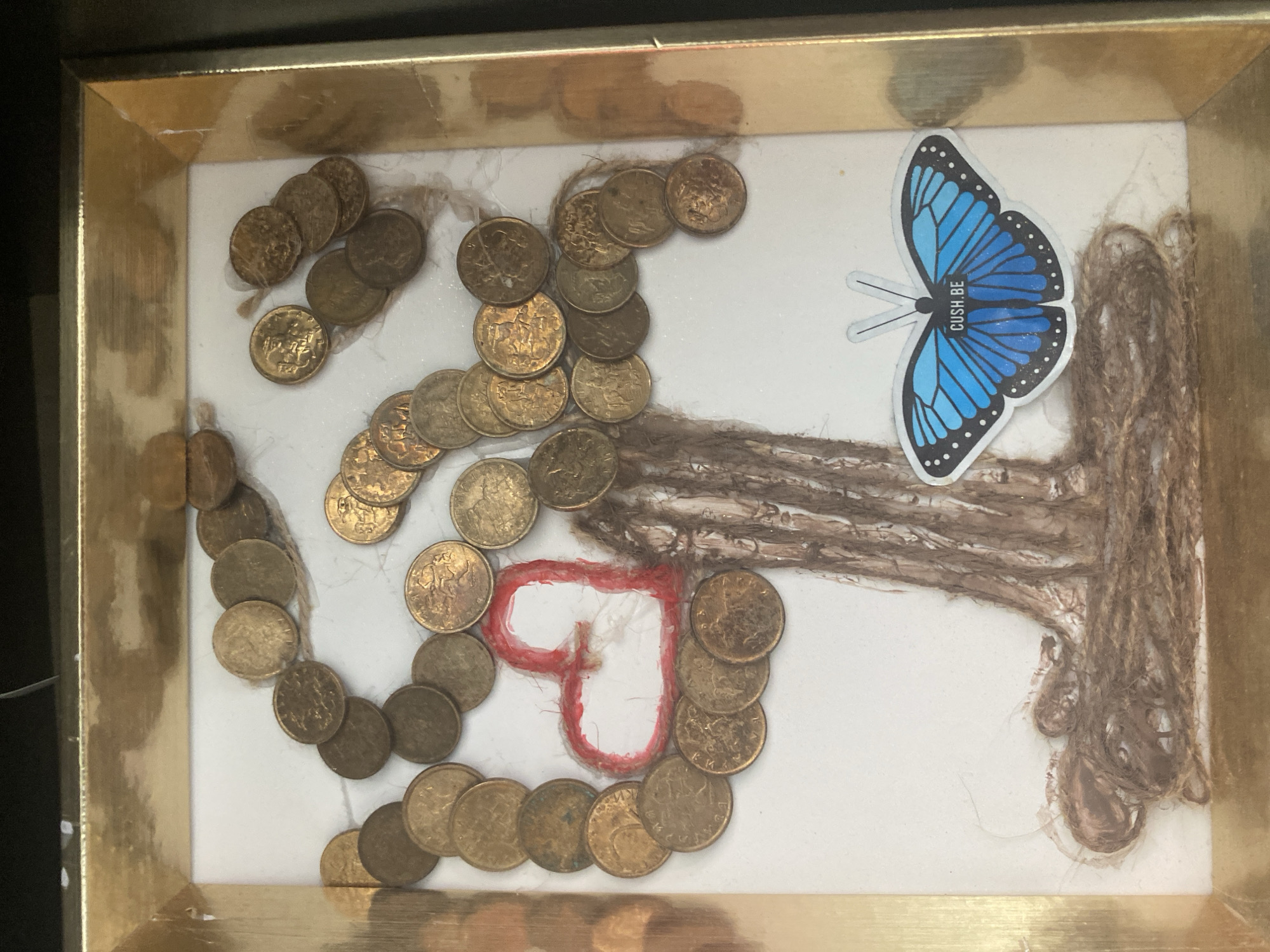 The image shows a framed shadow box containing a handmade art piece. The art depicts a tree with a trunk made of braided brown twine, roots made of the same twine at the base, and branches formed by an arrangement of approximately 40-50 circular, gold-colored coins. A red yarn heart is positioned on the left side of the tree. A blue butterfly sticker with the text "CUSH.BE" is affixed to the lower right of the frame, on top of the twine roots. The frame itself is gold-colored and appears to be made of wood or plastic. The background of the shadow box is white. The image is taken from a slightly overhead angle. No specific location in Blagoevgrad, Bulgaria is identifiable from the image.