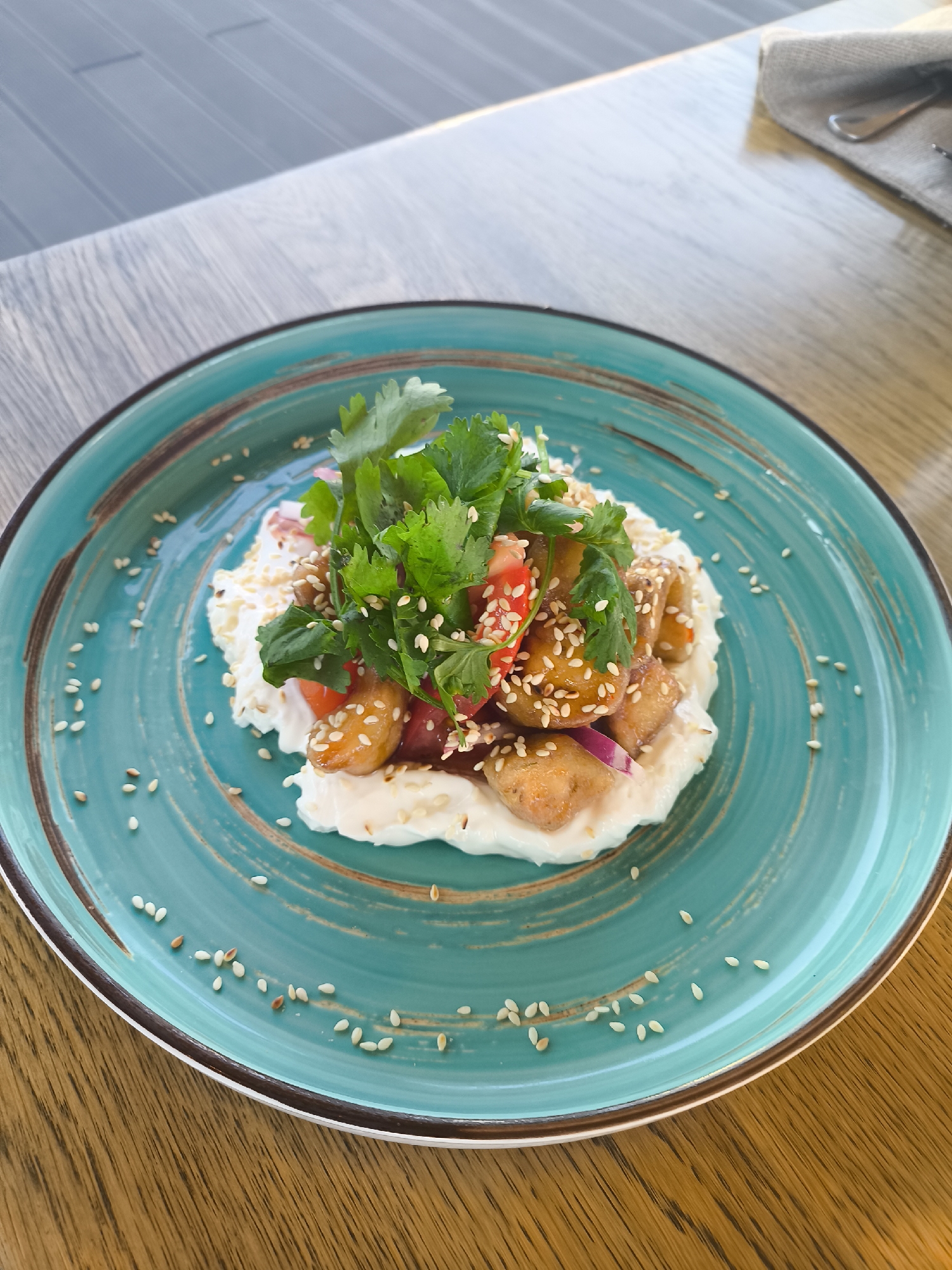 A vibrant, artfully arranged dish is captured in a close-up, eye-level shot, resting on a light brown wooden table. The central subject is a round, teal ceramic plate with a dark brown rim and subtle swirling patterns. Scattered across the plate's surface are numerous white sesame seeds.
At the center of the plate, the food is meticulously presented. A creamy, white base, resembling a thick sauce, yogurt, or labneh, forms a bed for the main components. Piled on top are golden-brown, bite-sized pieces, likely fried or sautéed protein such as tofu or chicken, many of which are generously coated with white sesame seeds. Interspersed among these pieces are finely chopped red elements, possibly tomatoes or red bell pepper, along with thin slivers of red onion. The dish is crowned with a fresh, verdant bunch of cilantro, adding a pop of green and a herbaceous garnish.
The wooden table has a visible grain, suggesting a natural material. In the out-of-focus background, hints of an indoor setting are visible, including more of the wooden table surface and a blurred element that appears to be a grey napkin with cutlery partially visible on the right side. The overall lighting is bright and natural, indicating it is likely daytime. There is no visible text in the image. The scene depicts a meal prepared for consumption, presented in an aesthetically pleasing manner.