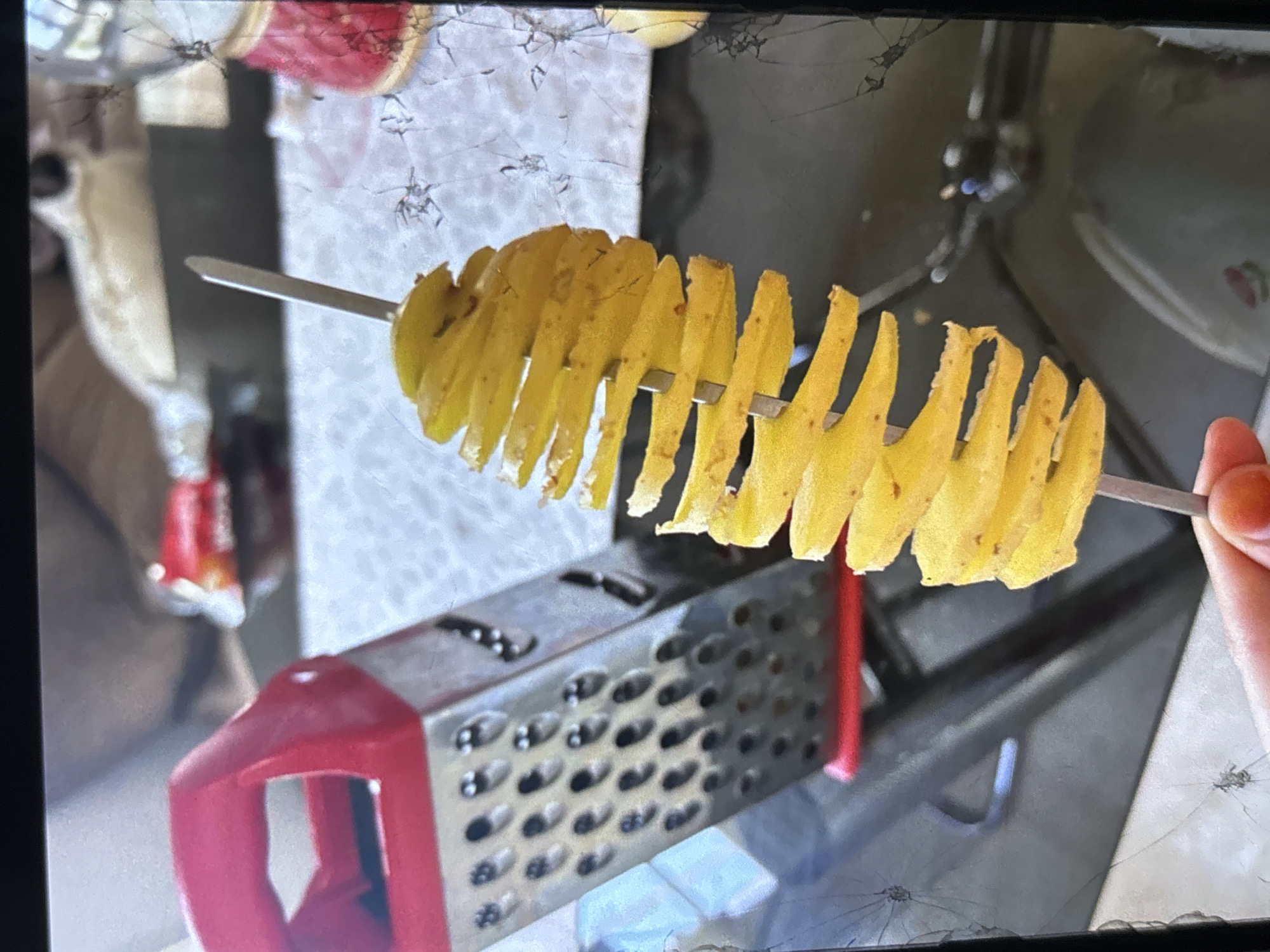 An overhead shot shows a hand holding a spiral-cut potato on a skewer. The potato is yellow and appears to be cooked. In the background, a red and silver grater is visible. The scene is set indoors, on a countertop with a white patterned surface. The image is slightly blurry and shows signs of damage, possibly from a cracked screen. The location of Orihuela, Spain, is not discernible from the visual information provided.