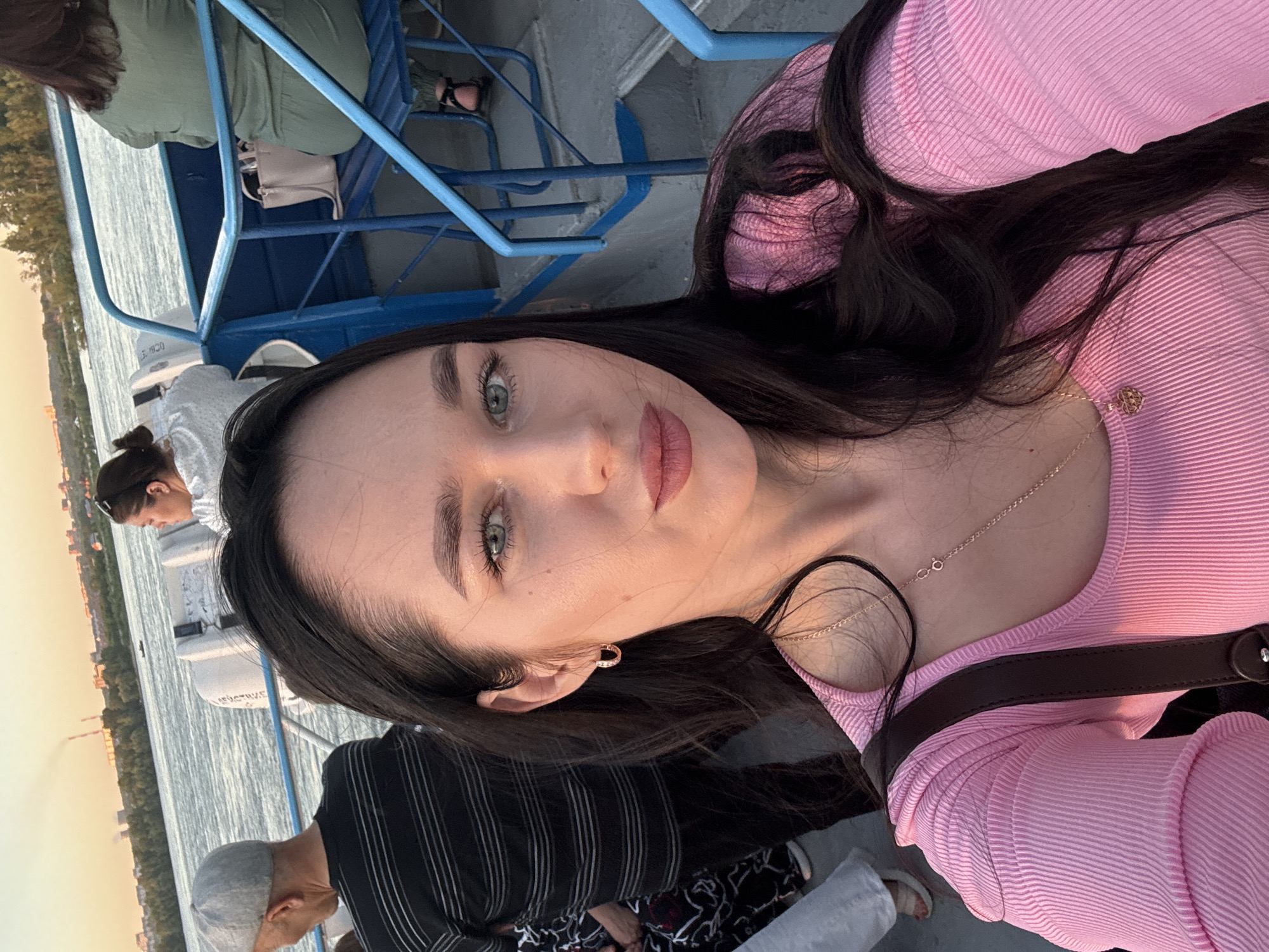 The image captures a selfie taken on a boat on a river, likely the Ob River, in Novosibirsk, Russia, during a sunset. The woman in the foreground, with long dark hair and green eyes, is wearing a pink ribbed long-sleeved shirt and a gold necklace. She is taking the photo with her right hand. Behind her, several other people are visible. To her left, an older man with a grey cap and a black and white striped shirt is facing away from the camera. Further back, a woman with dark hair tied in a bun is seated, wearing a white top. To the right of the woman in the foreground, another woman in a green outfit is seated on a blue bench with a beige bag. The boat appears to be a passenger vessel, with blue railings and seats. In the background, the Novosibirsk skyline is visible across the river, with buildings and industrial structures, and smoke rising from a chimney, indicating a city setting. The water is calm, reflecting the warm hues of the sunset. The overall atmosphere suggests a leisurely boat trip at dusk.