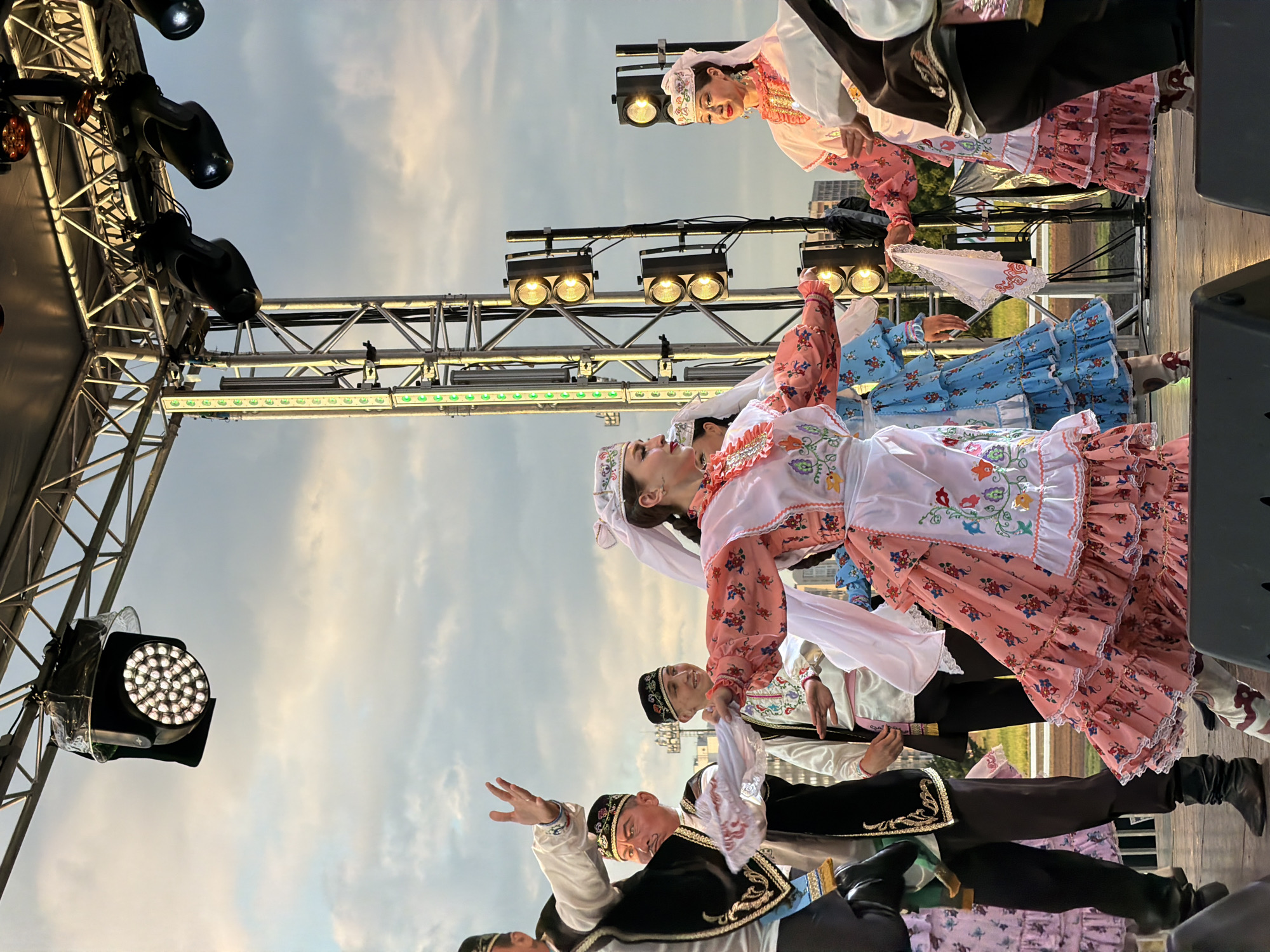 A group of people in traditional attire are performing on a stage. The performers include several women in ornate, long dresses and head coverings, and a few men in embroidered vests and hats. They appear to be dancing, with some holding scarves. The stage is set up outdoors, with a metal truss structure overhead supporting multiple lighting fixtures. The background shows a cloudy sky and hints of buildings and trees. The overall scene suggests a cultural performance or festival.