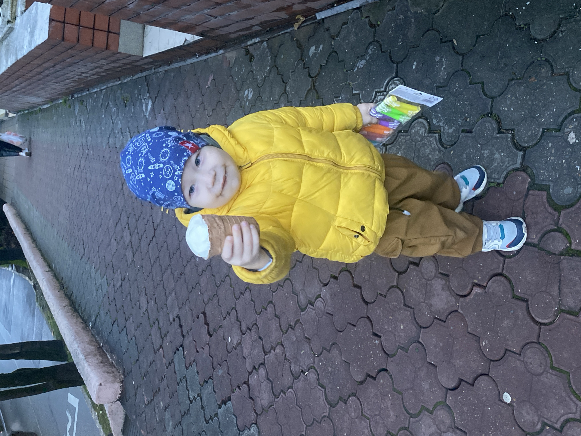 A young child, approximately a toddler, stands on a paved sidewalk in Sochi, Russia, facing the camera with a slight smile. The child is dressed in a bright yellow padded jacket, brown cargo-style pants, a blue beanie patterned with white and red celestial and textual designs, and light-colored athletic shoes with blue and dark grey accents. The child extends their right arm towards the viewer, holding an ice cream cone containing a single scoop of white ice cream. A small amount of white substance is visible on the child's chin. In their left hand, the child grasps a clear plastic package containing multiple colorful items, possibly markers or small toys. The sidewalk is constructed from dark grey and reddish-brown interlocking pavers. To the child's left, a light-colored curb separates the sidewalk from a street where a partial white line is visible on the asphalt. Several tree trunks line the far side of the street. To the child's right, a brick wall constructed from reddish-brown bricks rises, with a grey concrete base. Further down the sidewalk in the background, another individual is partially visible, walking away from the camera and carrying a white plastic bag.