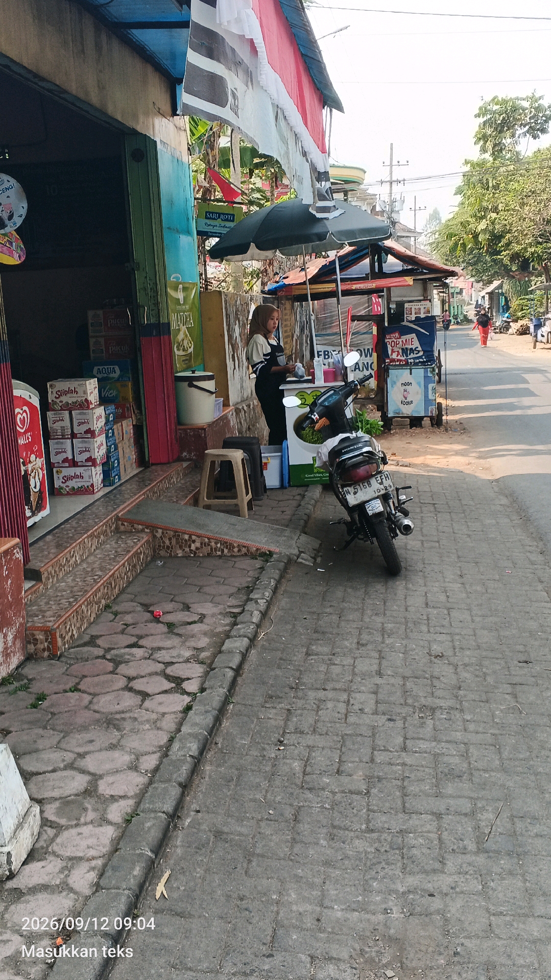 A street-level, eye-level, full outdoor shot depicts a sidewalk and the exterior of a shop in Tumpang, Indonesia. The shop, on the left, has a blue and green facade with a dark doorway. Boxes of products are stacked on the steps leading up to the shop. A woman wearing a hijab and an apron stands behind a food cart under a black umbrella, to the right of the shop. A motorcycle is parked on the sidewalk in front of the cart. The sidewalk is paved with interlocking bricks and has a cobblestone section on the left. Trees and other shops are visible in the background along the street. A man in an orange shirt walks in the distance on the right side of the street. The sky is clear and bright.