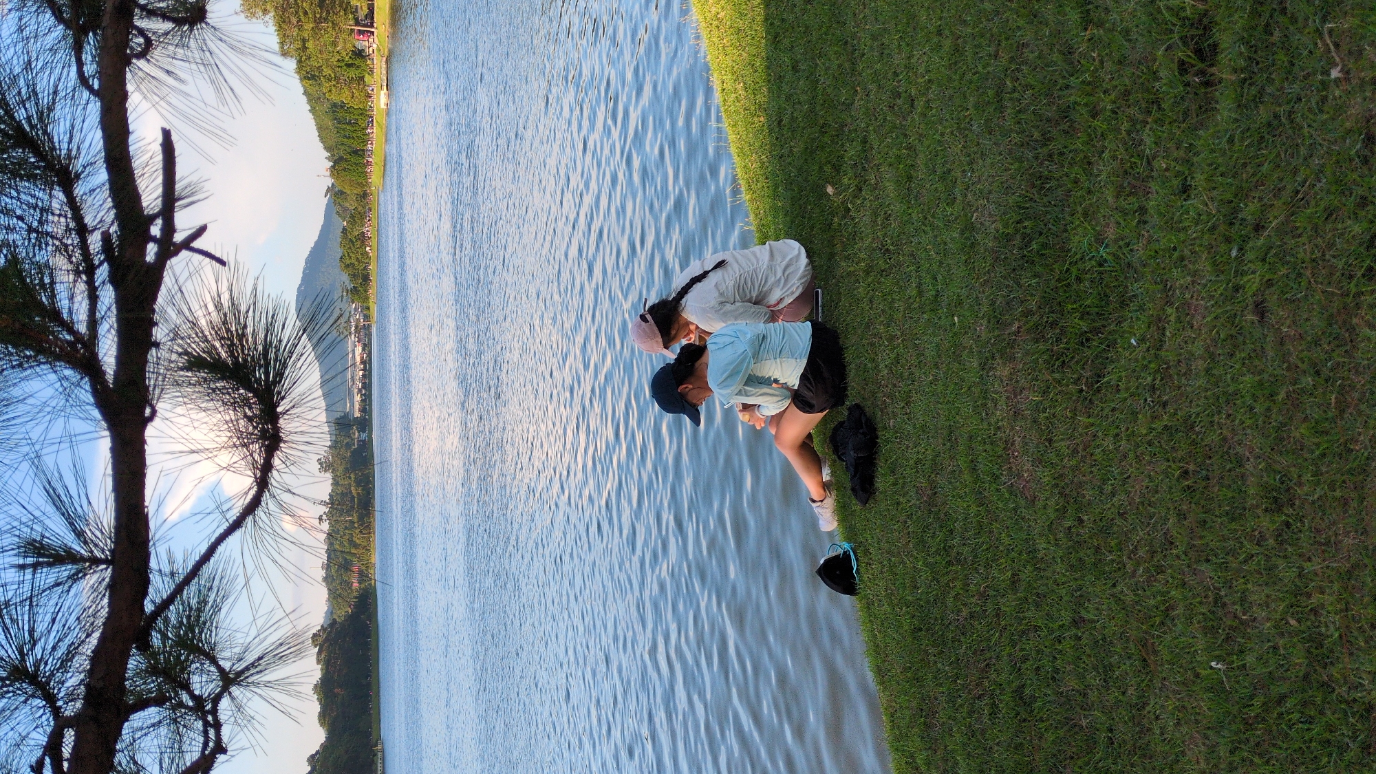 Two young females are sitting on a grassy bank beside a calm body of water, likely a lake or river, in Da Lat, Vietnam. The individual on the left is wearing a light blue long-sleeved shirt, black shorts, a dark blue baseball cap, and white sneakers. She is looking down at something in her hands. The person on the right is wearing a white long-sleeved shirt and light-colored shorts, with a pink baseball cap and her hair in a braid. She is also looking down. Between them on the grass are two dark-colored hats and a dark piece of clothing. The water reflects the sky, which is partly cloudy with visible sunlight. In the background, there is a shoreline with trees, and further in the distance, hills and mountains are visible, with some buildings suggesting a town or city. The foreground is dominated by the green grass. The scene suggests a peaceful, outdoor setting.