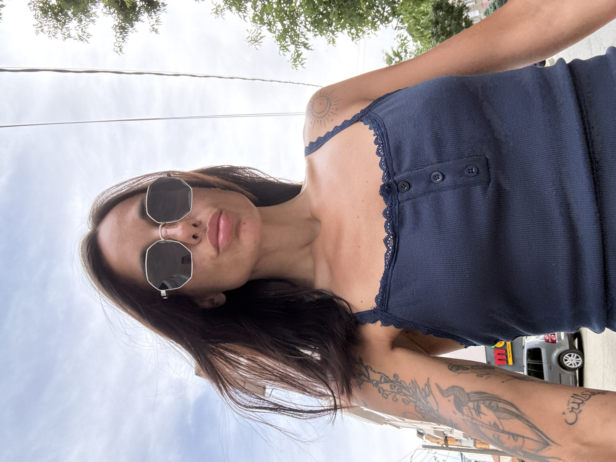 A low-angle, close-up, selfie-style shot features a woman with dark hair wearing a dark blue tank top and octagonal sunglasses. She is looking directly at the camera with her lips slightly pursed. She has tattoos on her left arm, including a detailed rendering of a face with horns and a bird with branches, and Arabic script. A sun and crescent moon tattoo is visible on her right shoulder. The background shows a cloudy sky with some trees and buildings on the left, and more trees on the right. A silver minivan is parked in the background, and a building with a red sign that says "M" is partially visible. The image appears to have been taken outdoors in a city environment, potentially on a street in Pleven, Bulgaria, given the architectural style of the buildings and the presence of a minivan typical of the region. The lighting suggests it is daytime.