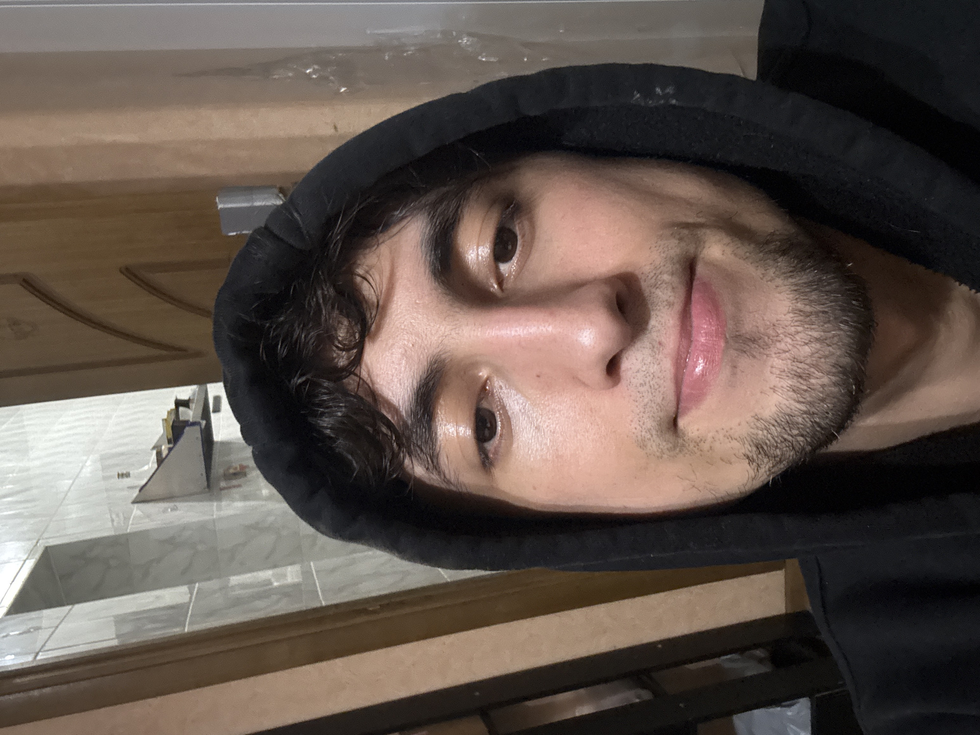 A close-up selfie shows a young man wearing a black hooded sweatshirt. His face is partially obscured by the hood. He has dark, curly hair, brown eyes, and a light beard. His skin appears slightly shiny, possibly from sweat or oil. The background is out of focus but suggests an indoor environment with tiled walls and wooden elements. The lighting is dim, with a warm tone.