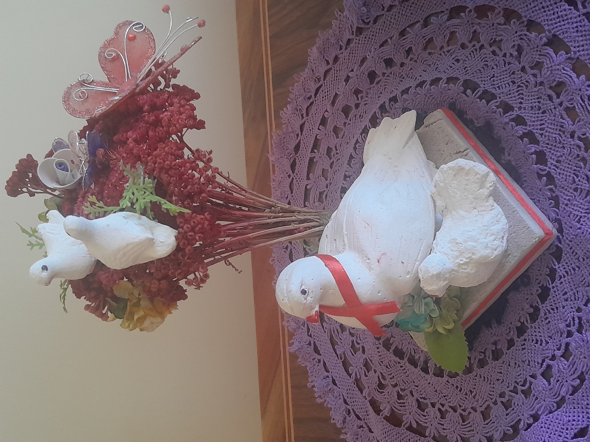 An indoor still life displays a ceramic figurine of a white dove with a red ribbon around its neck, perched on a base with a smaller, unfurled dove next to it. The figurine rests on a purple crocheted doily, which is placed on a wooden surface. In the background, a vase holds a dried floral arrangement featuring red berries and foliage, with a pink decorative butterfly and a white rose ornament protruding from it. Two smaller white ceramic doves are nestled within the floral arrangement. The lighting suggests the scene is illuminated by ambient room light. The location is described as Unknown, Unknown.