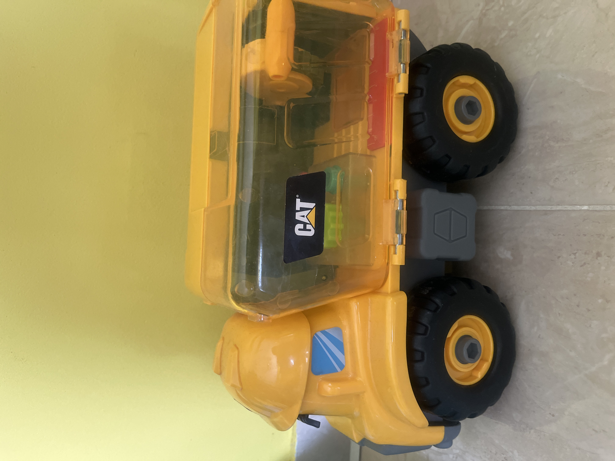 A bright yellow toy dump truck with a transparent orange lid is positioned on a tiled floor against a pale yellow wall. The truck features black wheels with yellow hubs and a "CAT" logo on its side. The lid of the truck appears to be hinged. There is no indication of the city of Onokivtsi, Ukraine, in the image.
