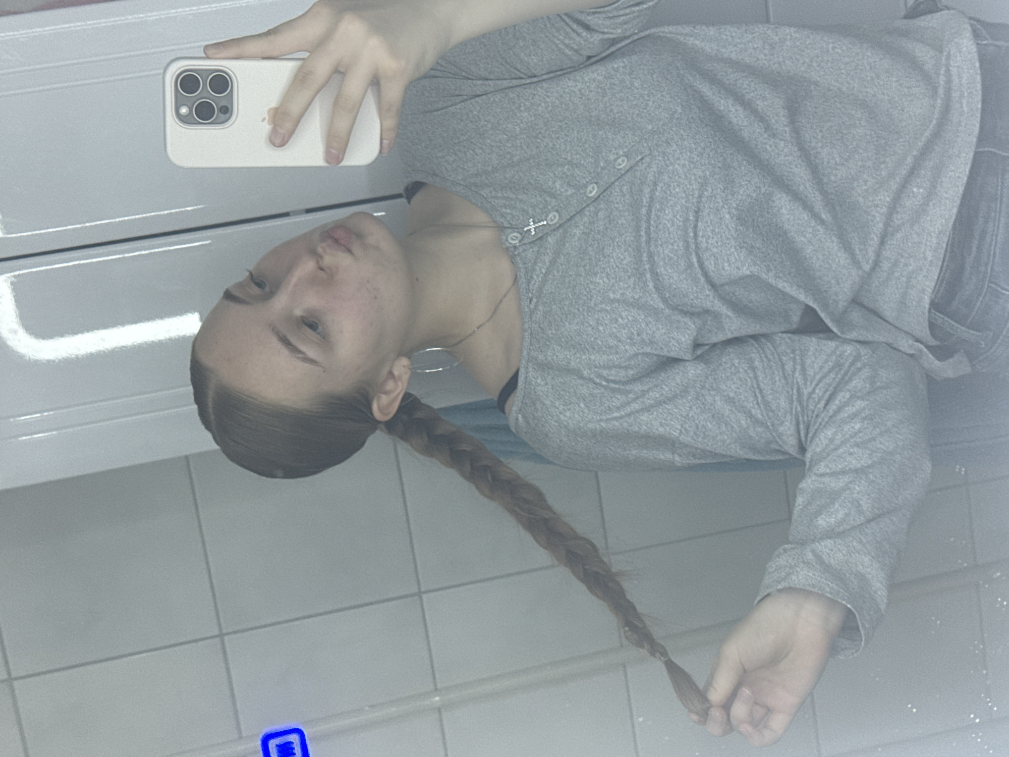 A young female with light brown hair styled in a neat, tight braid is captured in a mirror selfie. She is wearing a light gray long-sleeved Henley-style shirt with four white buttons and a visible black bra strap, large silver hoop earrings, and a thin silver chain necklace with a small cross pendant. Her lips are pursed, and she holds a cream-colored smartphone with a triple-lens camera array in her right hand, positioned to take the photo. Her left hand gently holds the end of her braid. The background, reflected in the mirror, consists of white rectangular tiled walls with dark gray grout lines, indicating an indoor setting, possibly a bathroom or kitchen. A prominent vertical reflection of an overhead light source is visible on the right side of the mirror, with a faint blue object reflected on the far left. The scene is set in an indoor location in Perm, Russia.