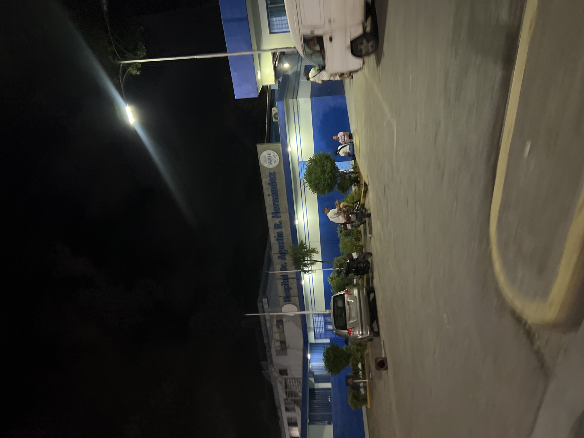 The image shows the exterior of Hospital Dr. Agustín R. Hernández in Juan Griego, Venezuela, at night. The hospital building is modern, with blue and white accents, and is illuminated by streetlights. Several vehicles, including a silver minivan and a white panel van, are parked in the lot. A few people are visible, some sitting on the ground near the entrance and others standing. The scene appears calm, with no overt signs of emergency or distress. The night sky is dark, with some visible clouds. The overall impression is of a functional hospital operating during nighttime hours.