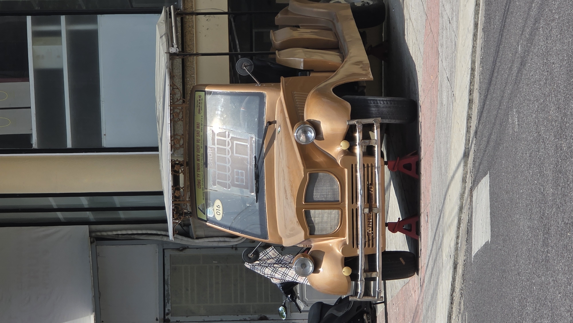 The image captures a distinctive gold-colored vehicle, possibly a custom-built tuk-tuk or motorized vehicle, parked beside a sidewalk in what appears to be Nha Trang, Vietnam. The vehicle is positioned on a concrete sidewalk bordering an asphalt street. The building beside the vehicle features glass windows with white frames and visible interior structures. Red vehicle stands are supporting the front of the vehicle. A parked motorbike partially obstructs the view of the front. The vehicle displays the number "016" and a logo on the windshield, suggesting a commercial or service-related function. The setting and design elements indicate an urban environment.