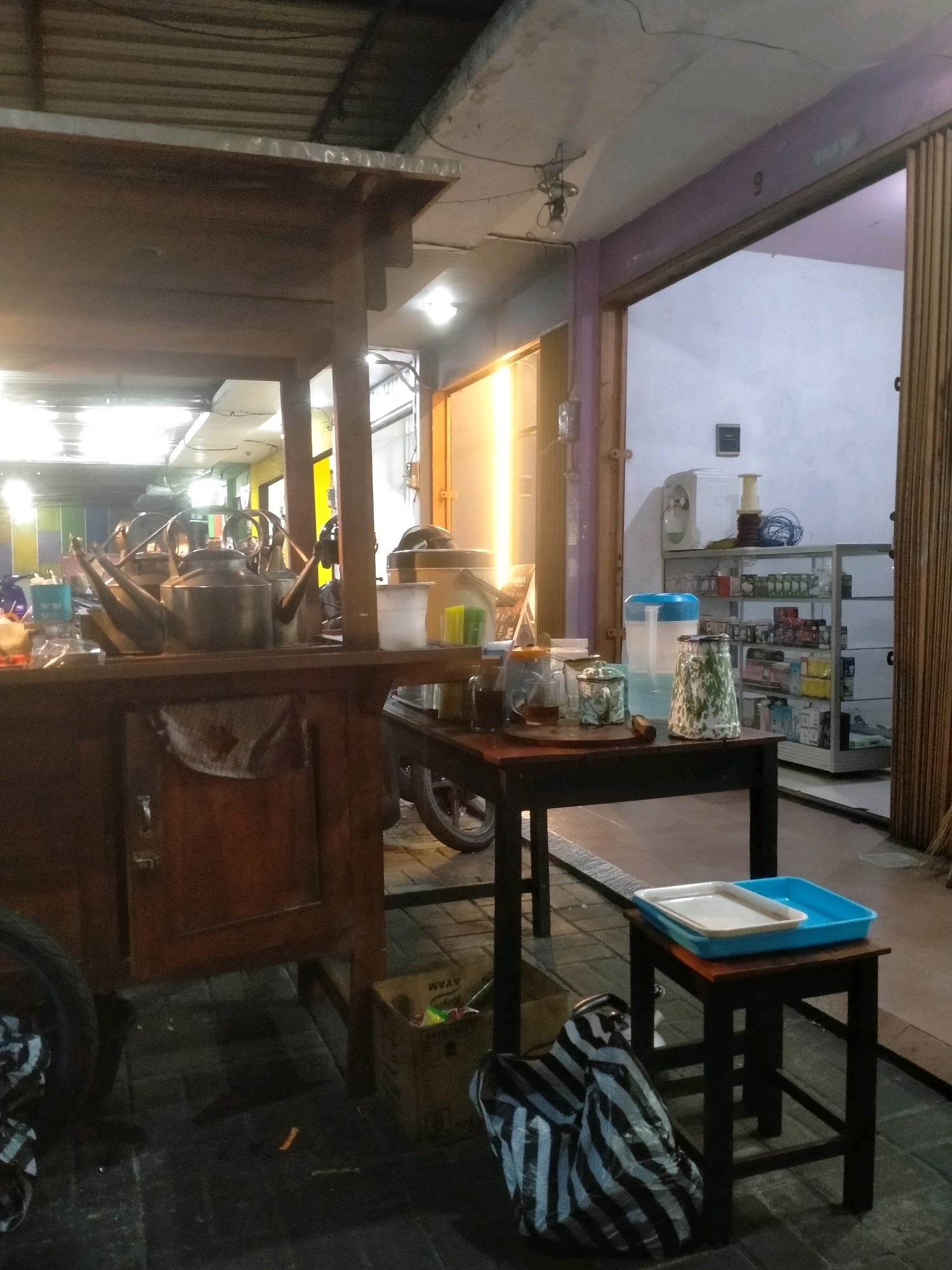 The image depicts a street-side food stall in Kasihan, Indonesia, at dusk. The stall features a wooden cart with a metal roof and a shelf displaying several metal teapots, suggesting it sells beverages. Adjacent to the cart, a dark wooden table holds various items, including a water dispenser, ceramic jars, and cups. To the right, a display shelf stocked with bottled goods and other products is visible within an open shopfront. The ground is paved, and a striped plastic bag is placed on a small stool in the foreground. The scene suggests a modest commercial operation, typical of local businesses in the area.