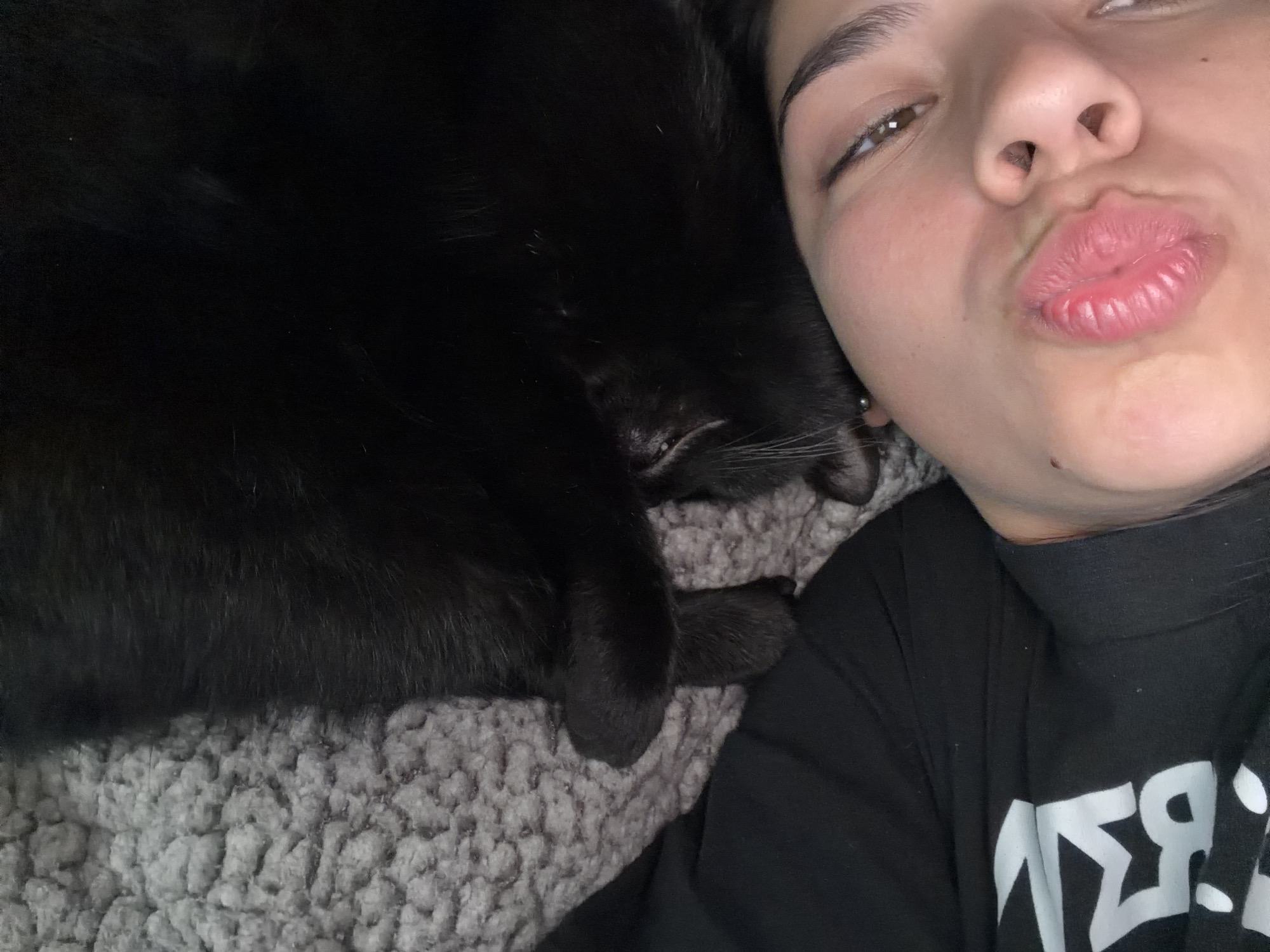 A selfie features a young woman with light skin and dark hair, pouting her lips toward the camera. Her right eye is visible, revealing a brown iris. She is wearing a black turtleneck shirt with white lettering. Nestled close to her is a black cat, lying on a gray, textured blanket. The cat's whiskers and paw are visible. The image lacks context to determine if the location is Gabrovo, Bulgaria.