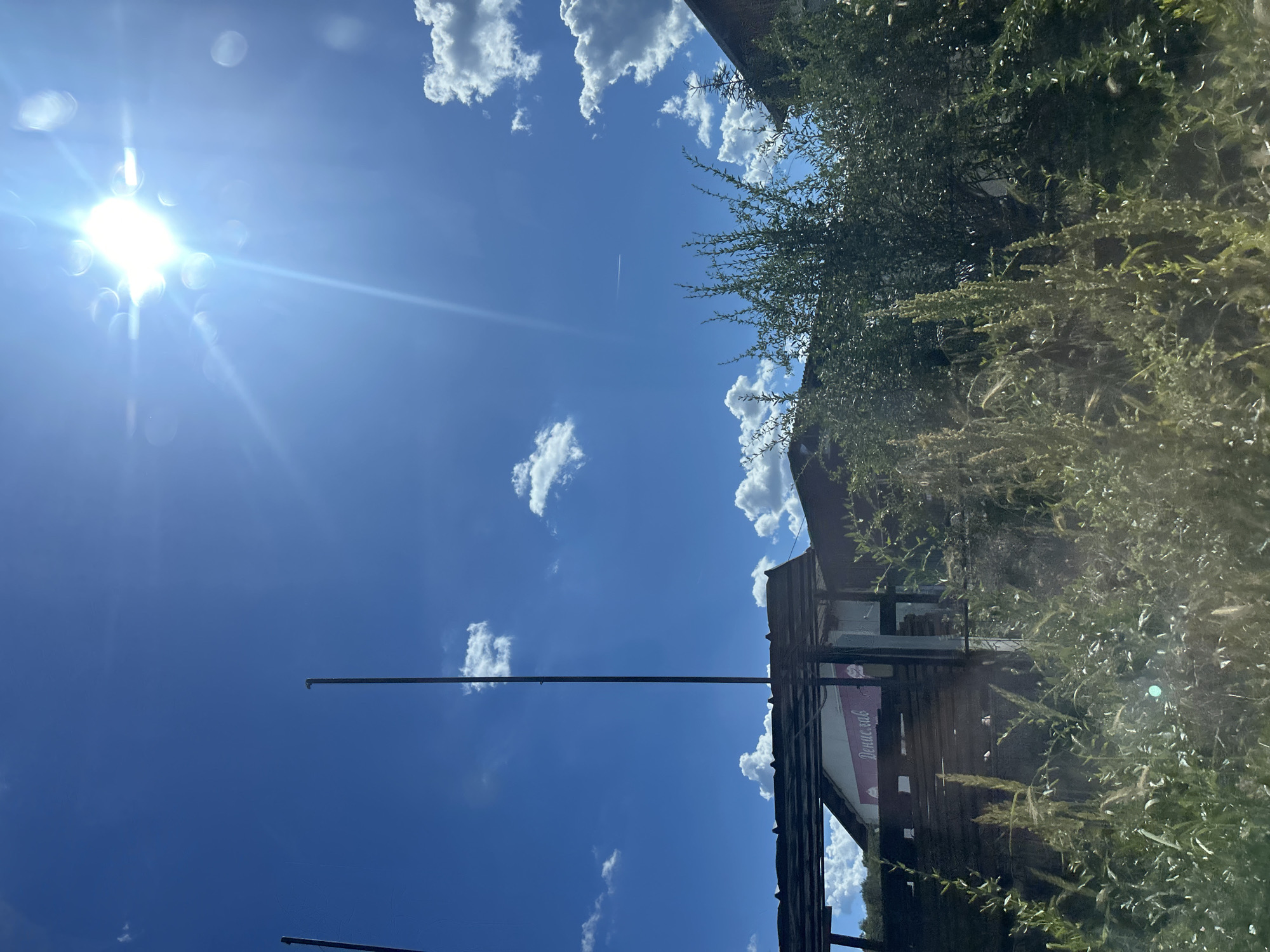 In Konstantinovo, Bulgaria, a bright sun illuminates a clear blue sky dotted with fluffy white clouds. Two tall, slender poles stand in the foreground, with one extending higher than the other. Beneath the sky, a rustic wooden structure with a corrugated roof is visible, partially obscured by lush green foliage. A sign with Cyrillic lettering, "Денислав," is affixed to the front of the building. The scene is bathed in sunlight, creating a warm and vibrant atmosphere.