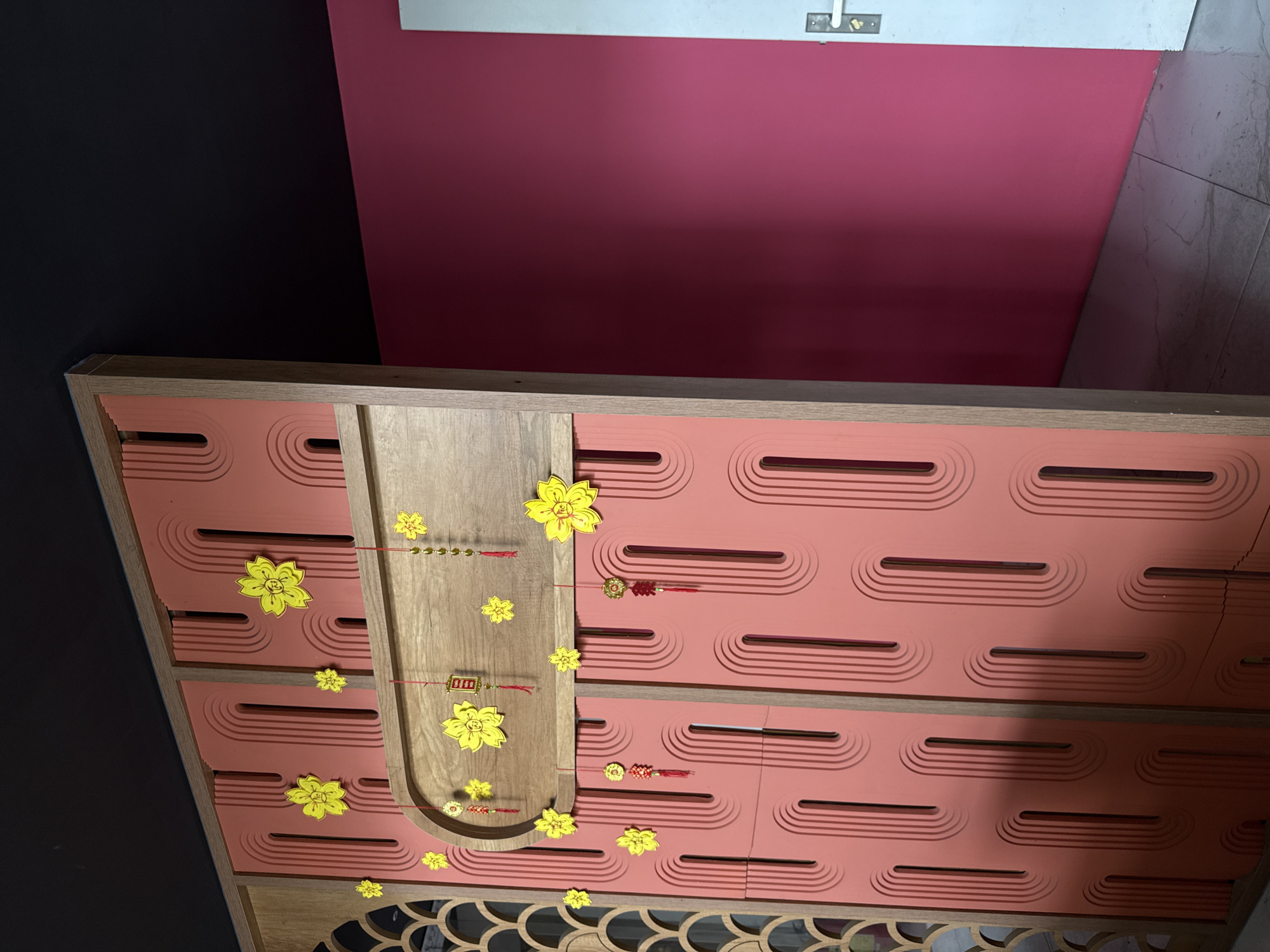 A close-up shot of a piece of furniture in Bac Nha Trang Ward, Vietnam. The furniture piece is made of light-colored wood and has a pinkish-red background with a pattern of horizontal, rounded lines. Yellow flower decorations are placed on the furniture, some attached with red string. The furniture's design includes a central wooden segment with a rounded arch and a decorative patterned base. The background consists of a dark wall on the left and a pinkish-red wall on the right, with a glimpse of a white shelf above the pink wall.