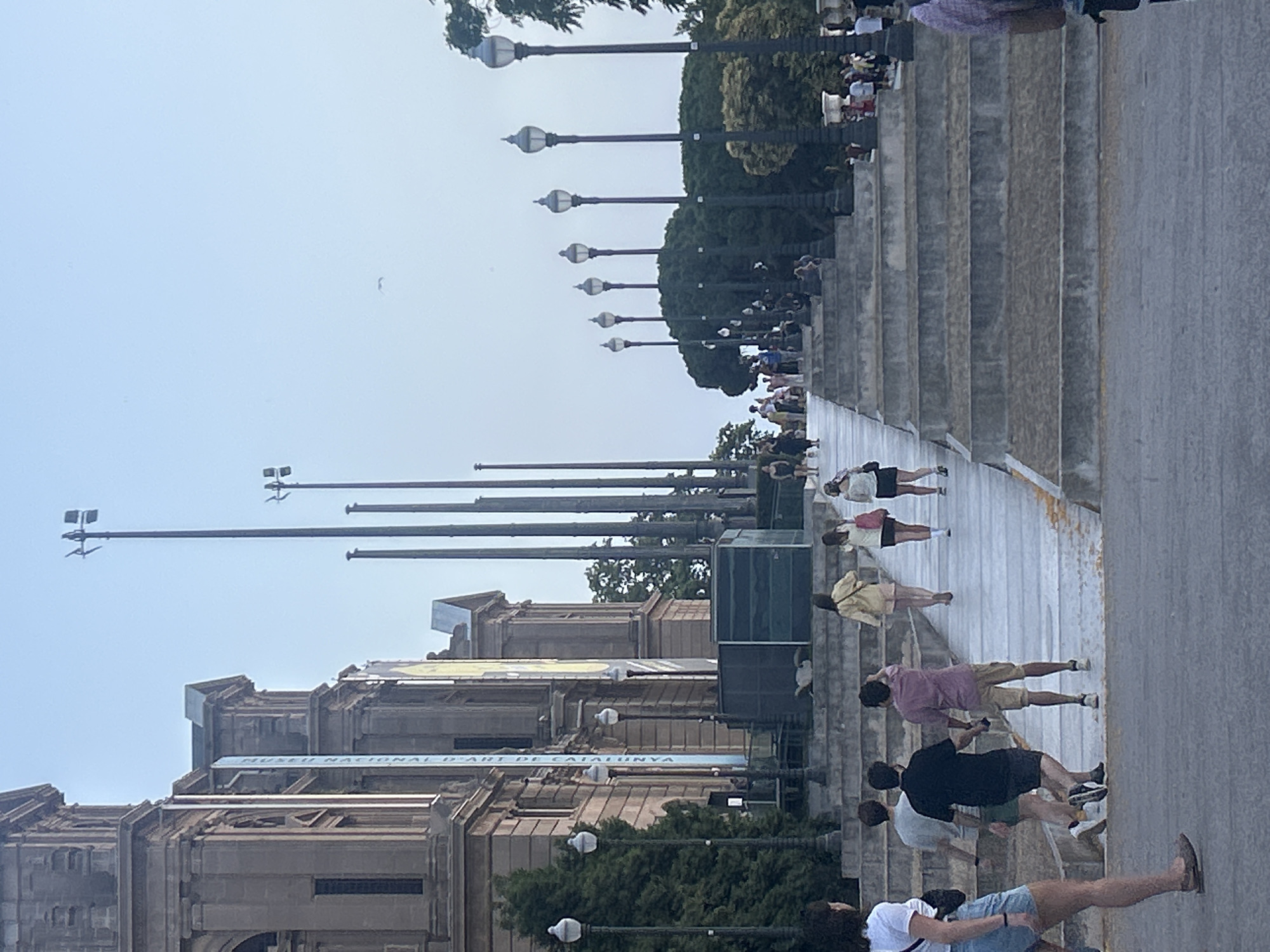 The image depicts the National Palace of Catalonia in Barcelona, Spain, a grand building with neoclassical architecture. In front of the palace, a wide set of stone stairs leads up towards the building. Several people are ascending and descending the stairs, dressed in casual summer attire. The atmosphere is clear and bright, suggesting it is daytime. Tall lampposts are strategically placed along the stairs, and trees are visible in the background. The overall scene conveys a sense of public space and tourism.