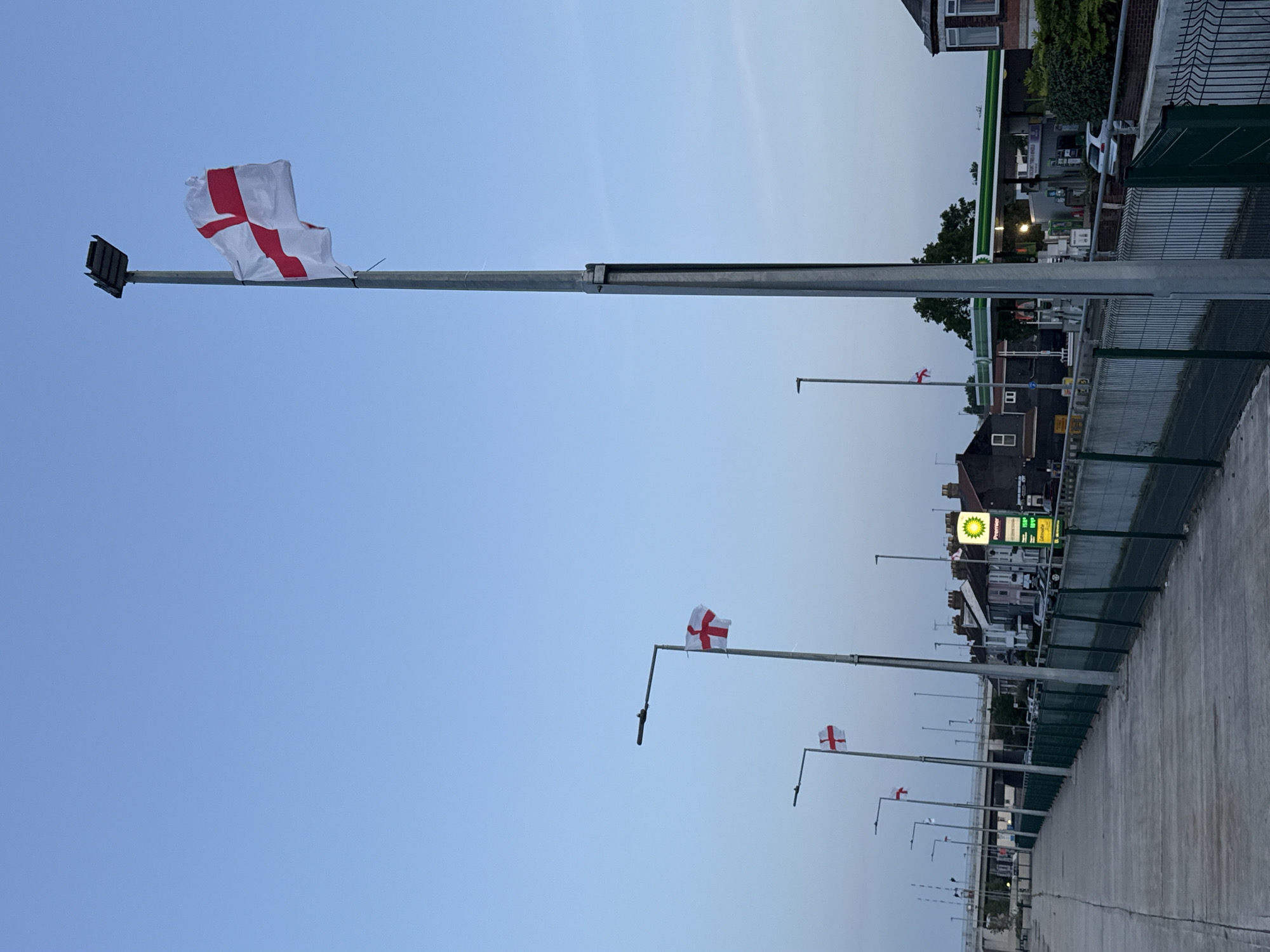 The image shows a row of flag poles lining a concrete walkway adjacent to a road. Each flag pole displays the flag of England: a white flag with a red cross. In the background, a BP gas station is visible, along with houses. The location is Great Yarmouth, United Kingdom. The sky is blue, suggesting either dawn or dusk. No people are visible in the image. The photograph is oriented vertically.