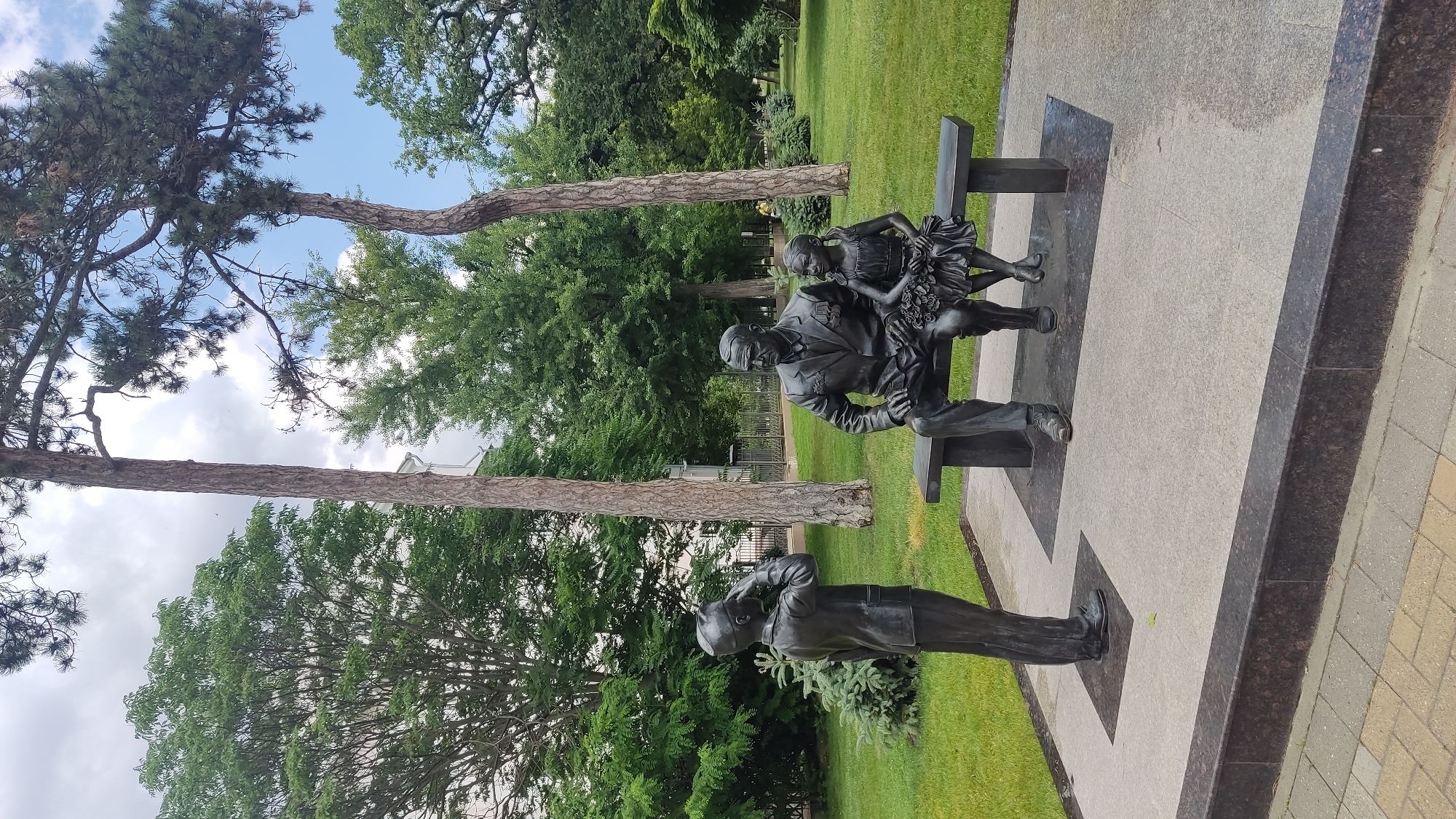 The image depicts a bronze sculpture in a park setting, possibly in the city of Krasnodar, Russia. The sculpture features three figures: an adult male seated on a bench, a young girl sitting next to him, and a young boy standing in front of them. The man is wearing a suit and medals, and the girl is in a dress. The boy is saluting. They are all rendered in dark bronze. The bench is also made of bronze and rests on a stone platform. The surrounding environment consists of green grass, trees, and a cloudy sky. The foreground shows a paved walkway with brick-like paving stones.