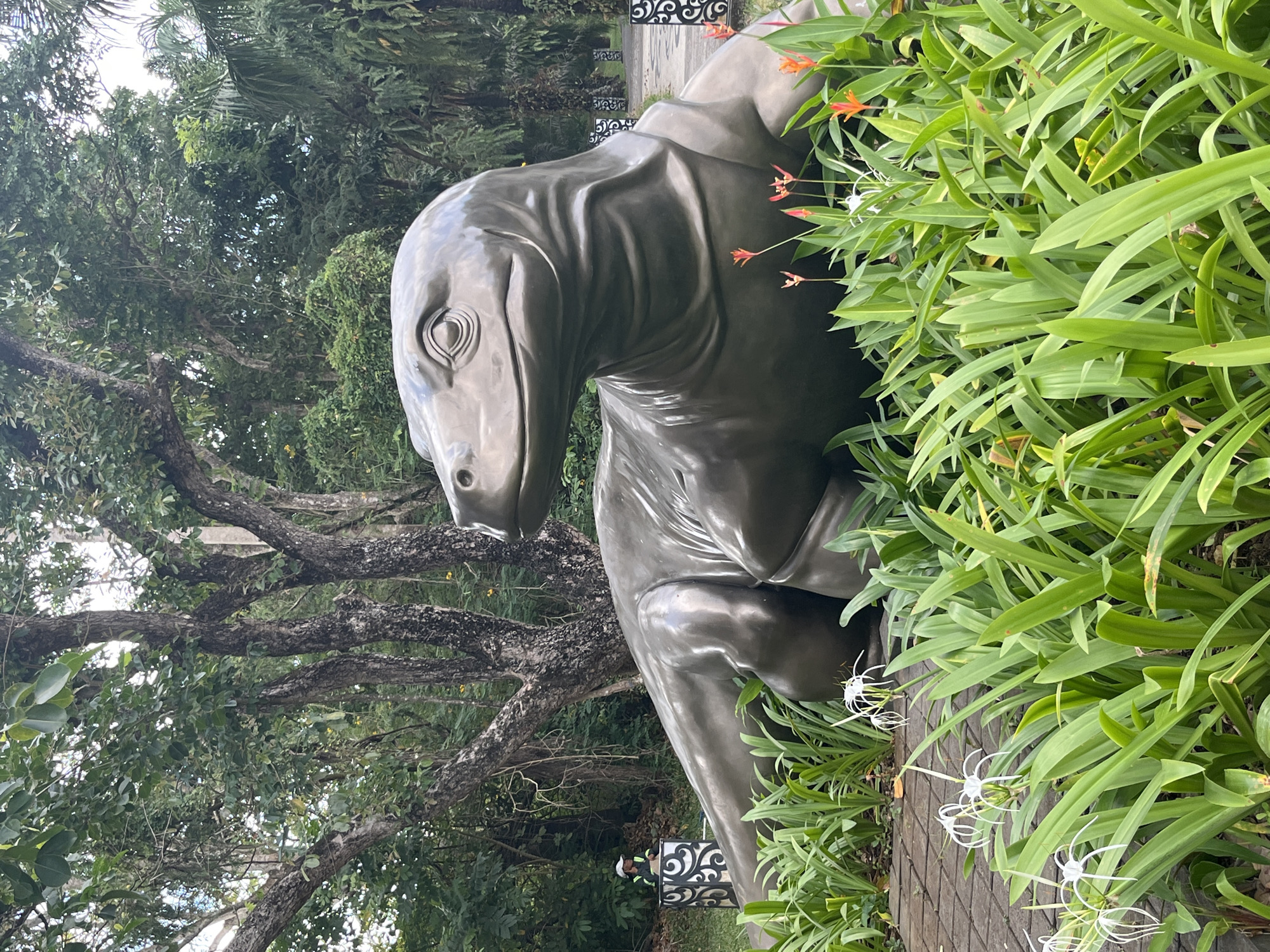 In Kuta Selatan, Indonesia, a life-sized bronze sculpture of a Komodo dragon rests in a lush garden. The sculpture is positioned on a paved walkway, surrounded by vibrant green foliage and delicate white flowers. In the background, tall trees with textured bark create a natural canopy, and a glimpse of a palm tree suggests a tropical setting. A person wearing a cap and a high-visibility vest stands behind a decorative patterned pillar, partially obscured by the vegetation. The Komodo dragon sculpture is the central focus, depicted with realistic detail and a lifelike posture, its head turned to the left, observing its surroundings. The overall scene evokes a sense of nature and art coexisting in a serene environment.