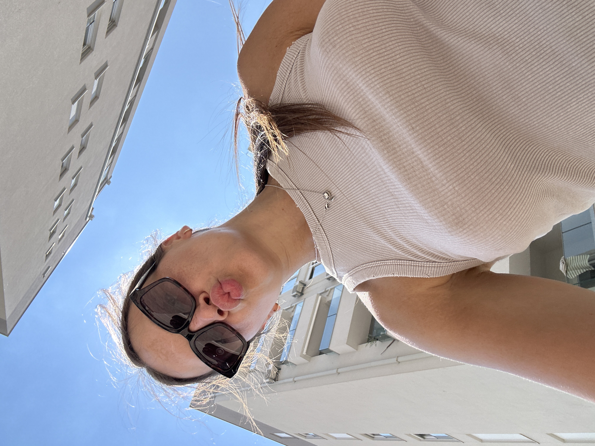 In Donji Pobori, Montenegro, a low-angle, wide shot captures a young woman taking a selfie against a bright blue sky. She is wearing large, dark sunglasses and a light-colored, ribbed tank top. Her lips are pursed, and her hair is blowing in the wind. Behind her, the modern architecture of buildings, one featuring a facade with diamond-shaped window cutouts, dominates the background, with balconies visible on the other. The lighting suggests it is a sunny day.