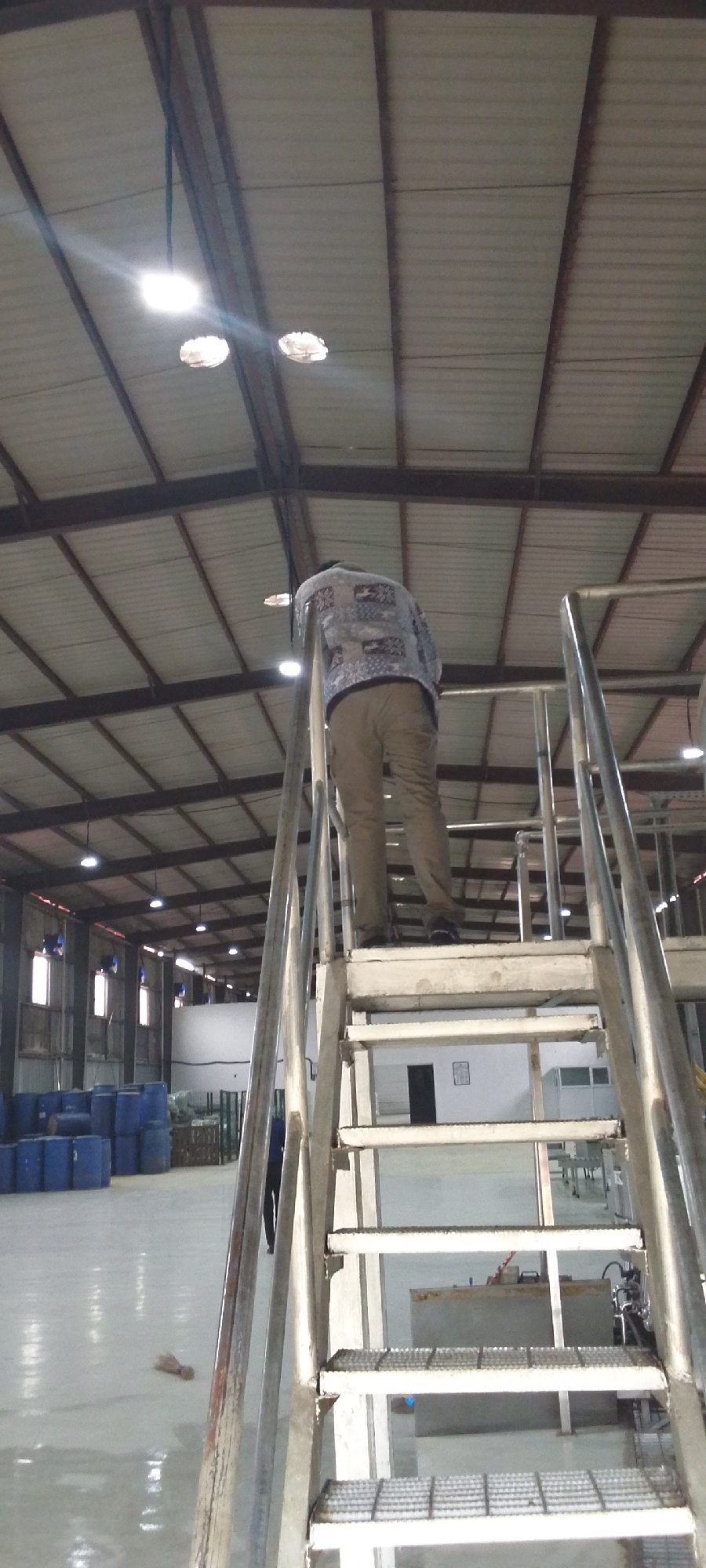 The photograph depicts the interior of what appears to be a large industrial warehouse. The perspective is from the base of a metal staircase, looking upwards towards a person standing on the stairs. This person is wearing a patterned shirt and khaki pants.
In the background, rows of blue barrels are visible on a polished concrete floor. Overhead, the warehouse has a high ceiling constructed of corrugated metal panels supported by a steel frame. Lighting fixtures are installed along the ceiling. A piece of what appears to be clothing is lying on the floor near the base of the stairs. A white container is present to the center left of the image.