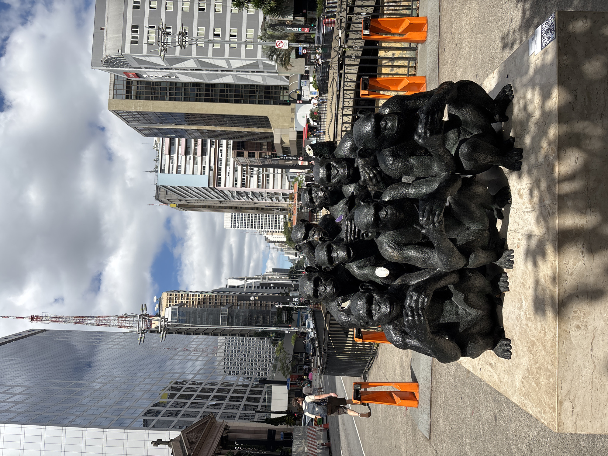 A street-level shot shows a sculpture of dark, clustered figures in São Paulo, Brazil. The sculpture rests on a marble base with orange barriers placed around it. A person is walking past. Buildings tower in the background, with a partly cloudy sky visible above. The street appears to be busy with traffic and pedestrians. A crane is visible in the distance atop a tall building.