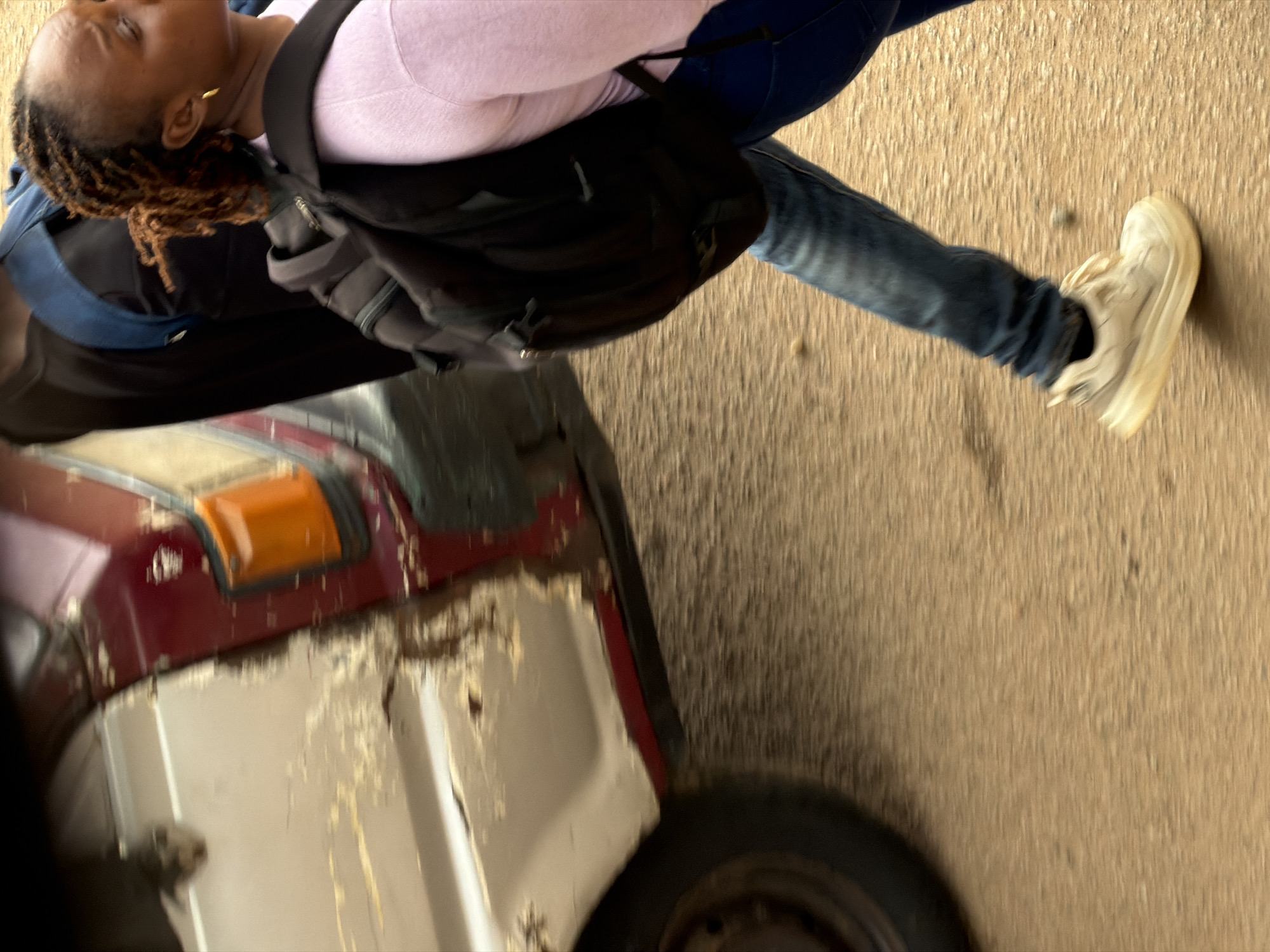 In Bukuru, Nigeria, a person with braided hair and a gold earring walks down a textured, sandy road. They are wearing a light pink long-sleeved shirt, blue jeans, and white sneakers, and carrying a black backpack. To the left, a weathered vehicle with a damaged white and red exterior is partially visible, its orange turn signal illuminated. The scene is captured with motion blur, suggesting movement.