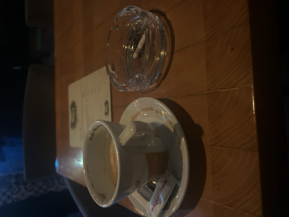 A table setting features an ashtray with a cigarette butt, a menu, and a cup on a saucer. The cup appears to contain a beverage. A small package of creamer and a spoon rest on the saucer. The table has a brown, wooden surface with visible seams. Lighting is dim, with darker areas to the right of the frame.