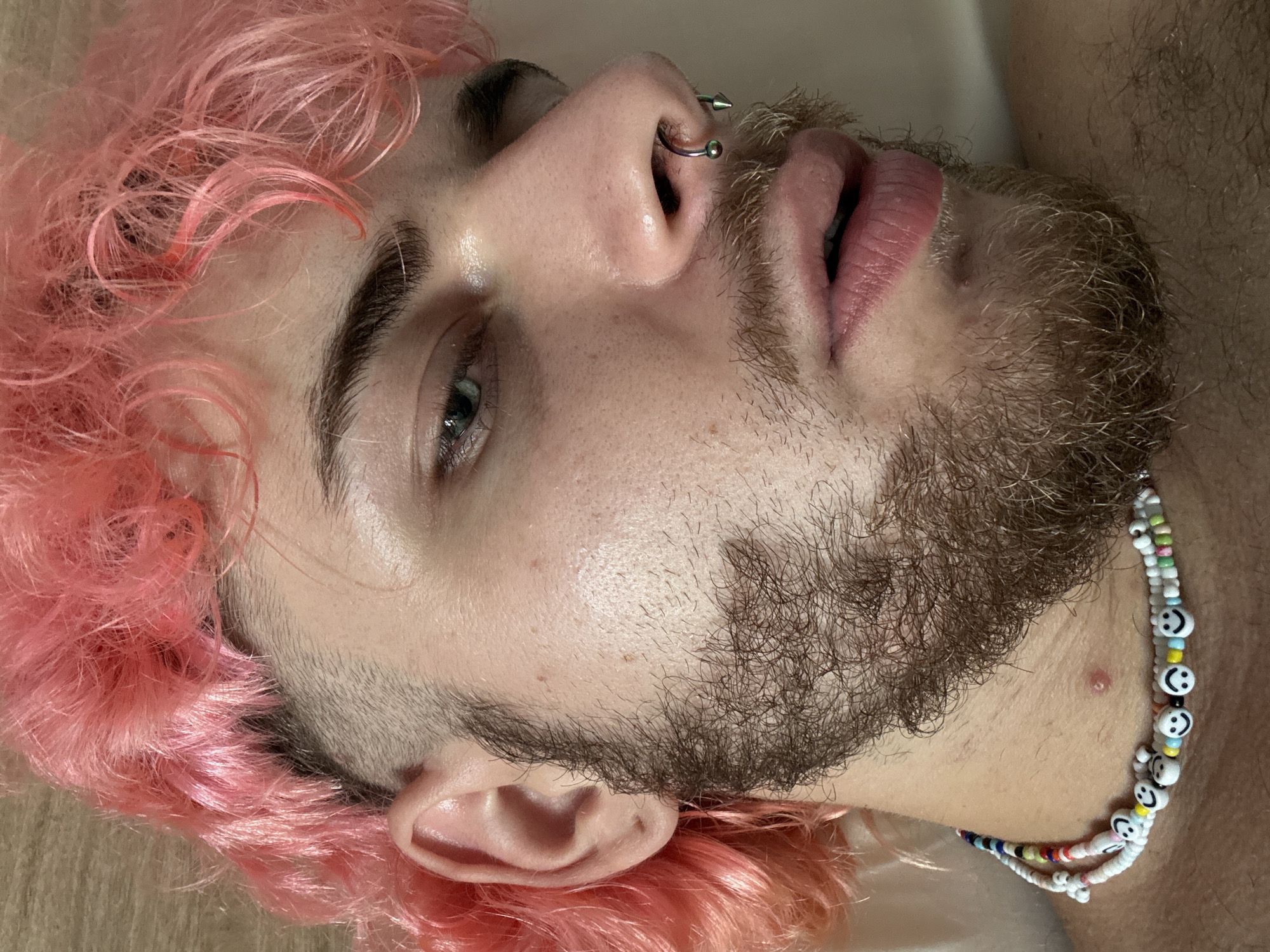 A close-up, slightly tilted image captures a person with pink hair and a brown beard. They have a nose piercing and are wearing a beaded necklace with smiley face designs. The person's skin appears fair. No information about the location of Đà Nẵng, Vietnam can be found in the image.