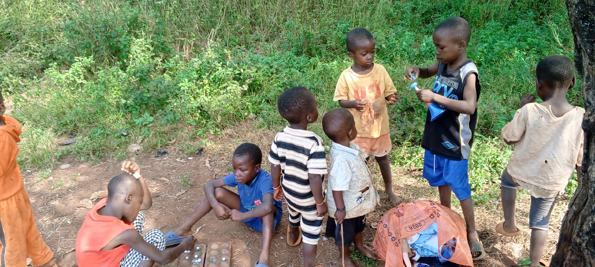 In Ngalula, Democratic Republic of the Congo, a group of African children are gathered in a dirt clearing near vegetation. One child in an orange jumpsuit stands at the left edge. Another, wearing an orange shirt and checkered pants, sits on the ground playing a game with marbles and a wooden board, along with a child in a blue shirt. Nearby, another child in a striped shirt and shorts stands watching. Three more children stand in a group, one in a yellow shirt, one in a black tank top and blue shorts holding a blue container, and one in a light-colored shirt. An orange bag full of items is on the ground. A tree trunk is visible on the right.