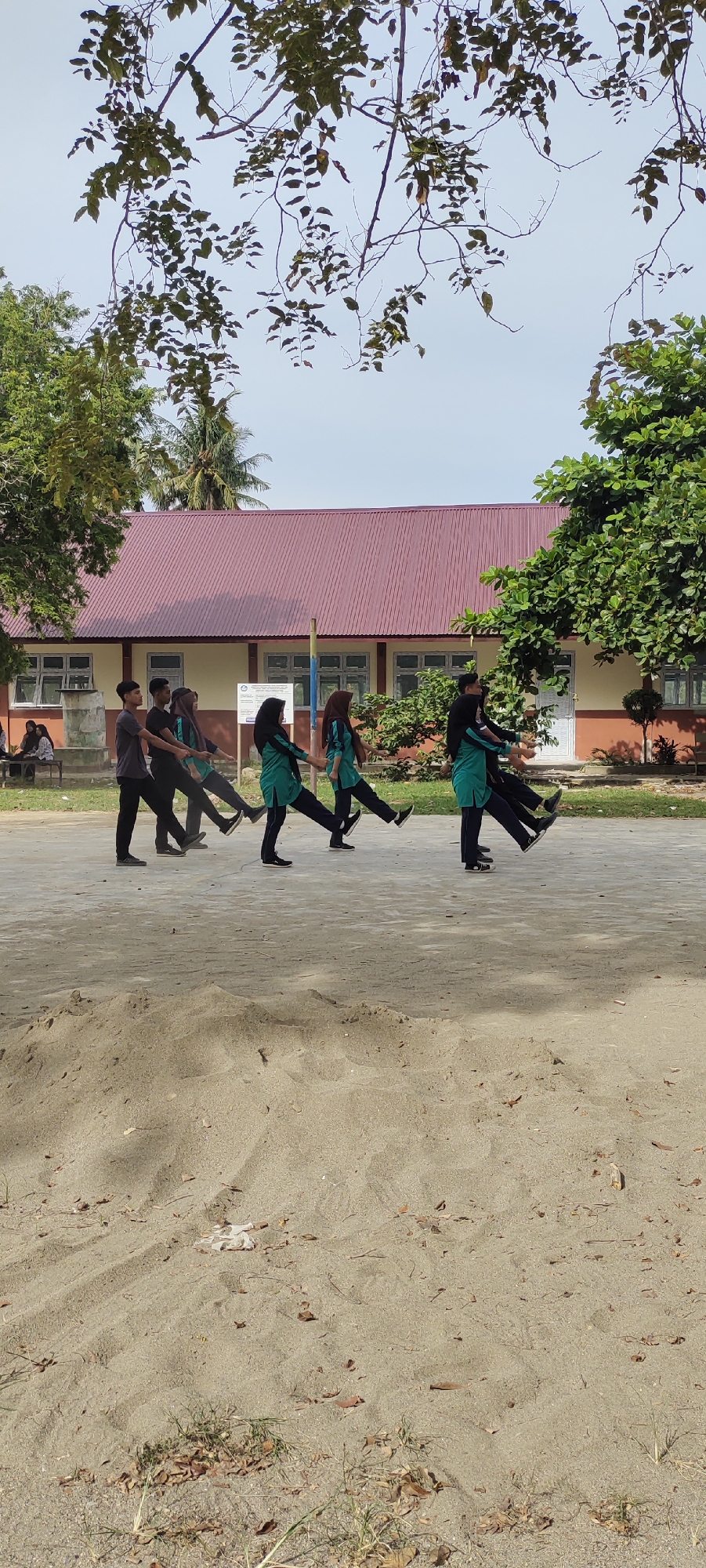 A group of seven young people are engaged in a marching drill on a sandy outdoor area in front of a two-story building with a red roof. The building appears to be a school or institutional facility, with several windows visible on the ground floor. Trees with green foliage are scattered around the building and the foreground, with one large tree on the right side of the frame casting a shadow. The ground is a mix of sand and some patches of dry grass and leaves.
The individuals are dressed in uniforms. The first three individuals from the left are wearing dark grey t-shirts and black pants, with two of them wearing headscarves. The next four individuals are wearing teal-colored shirts with long sleeves and black pants, also with headscarves. They are all in mid-stride, with their arms extended and hands clasped with the person in front of them. The drill appears to be a synchronized marching formation.
In the background, to the far left, a few people are sitting on a bench, and a water tank is visible. The sky is clear and light blue. The overall scene suggests an organized outdoor activity, possibly a school event or practice session, taking place in a tropical or subtropical climate. The location is specified as Langnibong, Indonesia.
