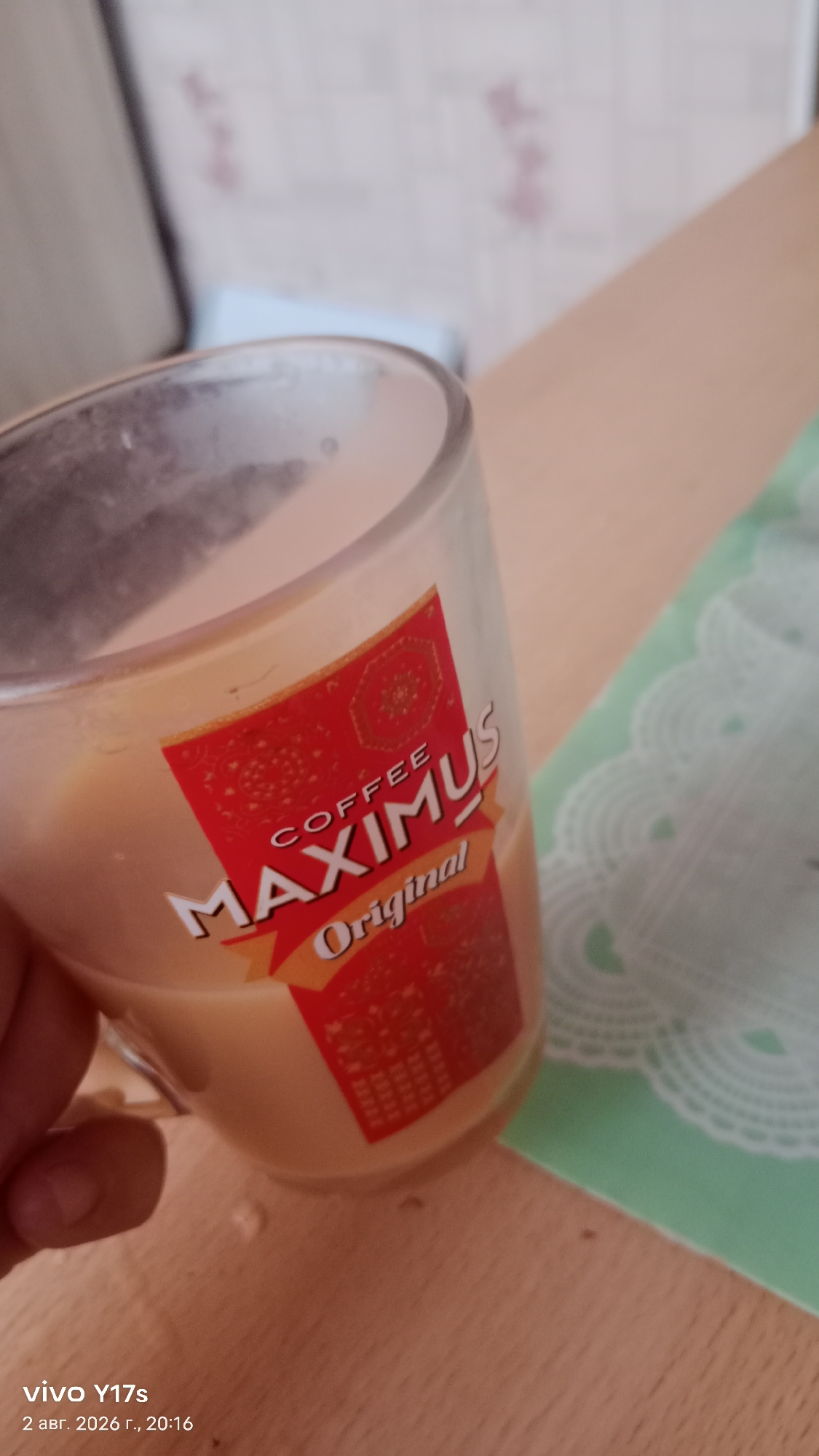 A glass mug filled with a light-colored beverage, possibly coffee, is held by a person's hand. The mug has a red label with the text "COFFEE MAXIMUS Original" printed in white and gold. The background is blurred but appears to be an indoor setting with a patterned wallpaper and a wooden table. A green doily or placemat is visible to the right of the mug. The timestamp in the lower left corner indicates the photo was taken on August 2, 2026, at 20:16, and the device used was a vivo Y17s. The location is specified as Rudny, Kazakhstan.