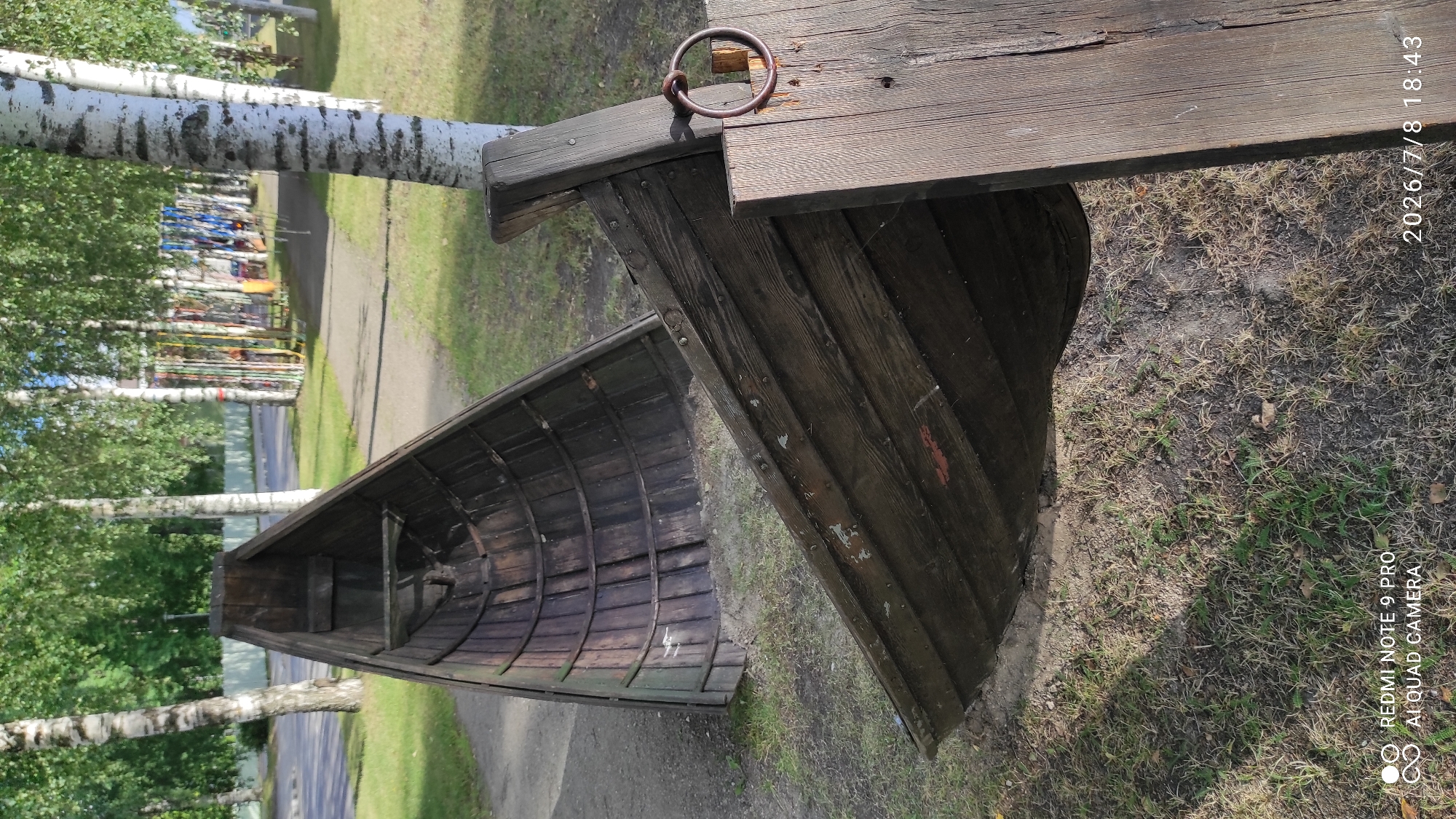 Two old wooden boats are positioned outdoors in a park-like setting in Iisalmi, Finland. One boat is in the foreground, appearing to be partly submerged in the ground and resting on a wooden plank. It is dark brown and weathered, showing signs of age and wear. The other boat is in the background, also wooden and dark brown, positioned at an angle on a path. The boats are surrounded by green grass and a path, with birch trees and other foliage in the background. In the far background, there are playground-like structures with colorful ropes and poles. The sky is overcast. The image includes a timestamp in the lower right corner indicating July 8, 2026, 18:43.