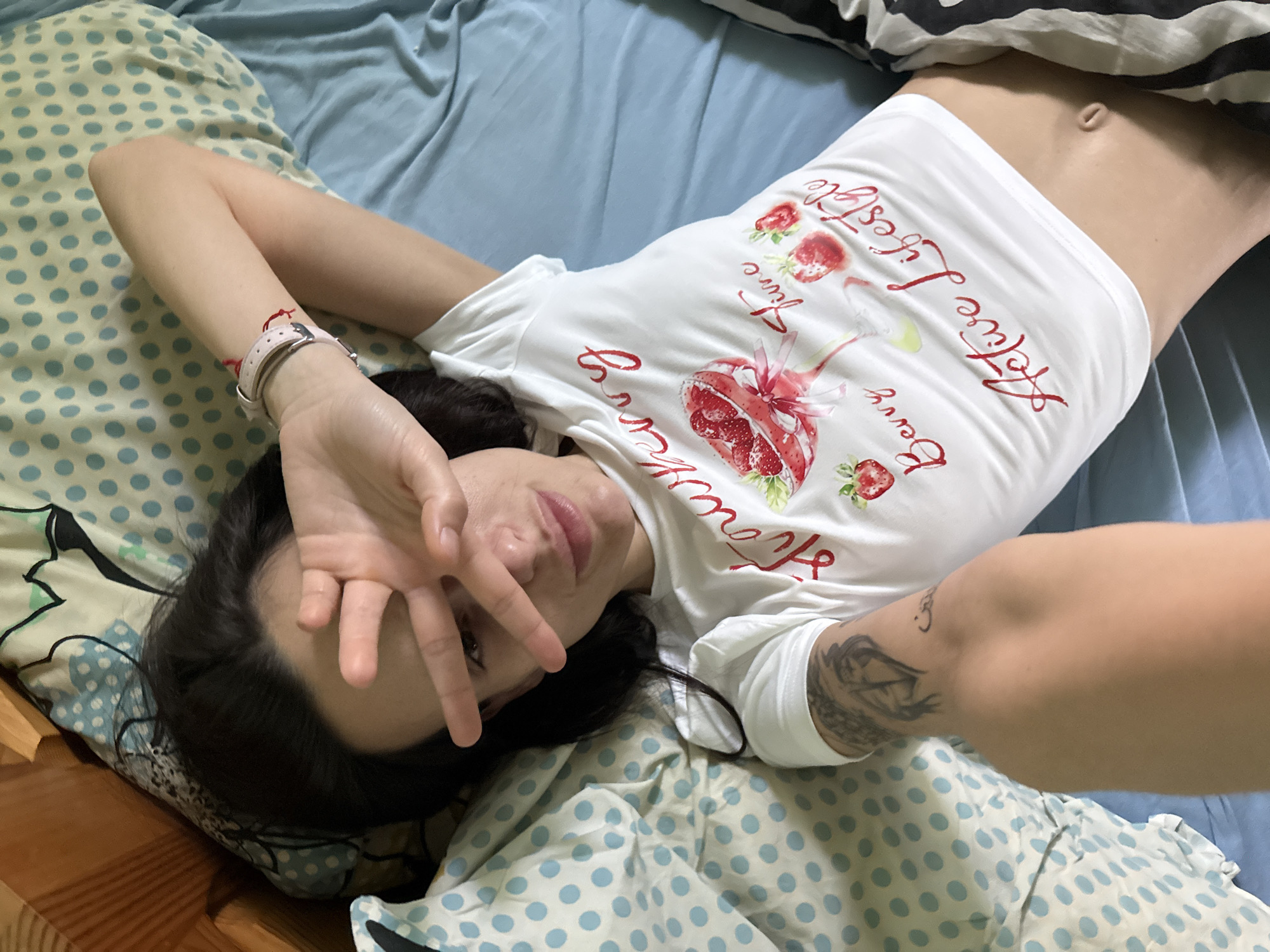 A female individual with dark hair is depicted lying supine on a bed in Pleven, Bulgaria. She is wearing a white short-sleeved crop top featuring red text and graphics that read "Strawberry Berry Active Lifestyle" and illustrate a strawberry cocktail glass. Her left hand (viewer's right) is raised, with fingers partially obscuring her face, specifically her eyes and forehead, in a "V" gesture. A light pink watch with a white strap and a red string bracelet are visible on her left wrist (viewer's right). A dark tattoo, appearing to depict a bird or creature head, is visible on her right upper arm (viewer's left), alongside script. Her navel is exposed below the crop top. The bedding consists of a light blue sheet on the right side and a light blue and white polka-dotted sheet or pillow on the left, with an additional yellowish polka-dotted pattern visible at the top left. A wooden bed frame element is partially visible on the far left. Black and white striped fabric is visible at the bottom right, covering the lower body.