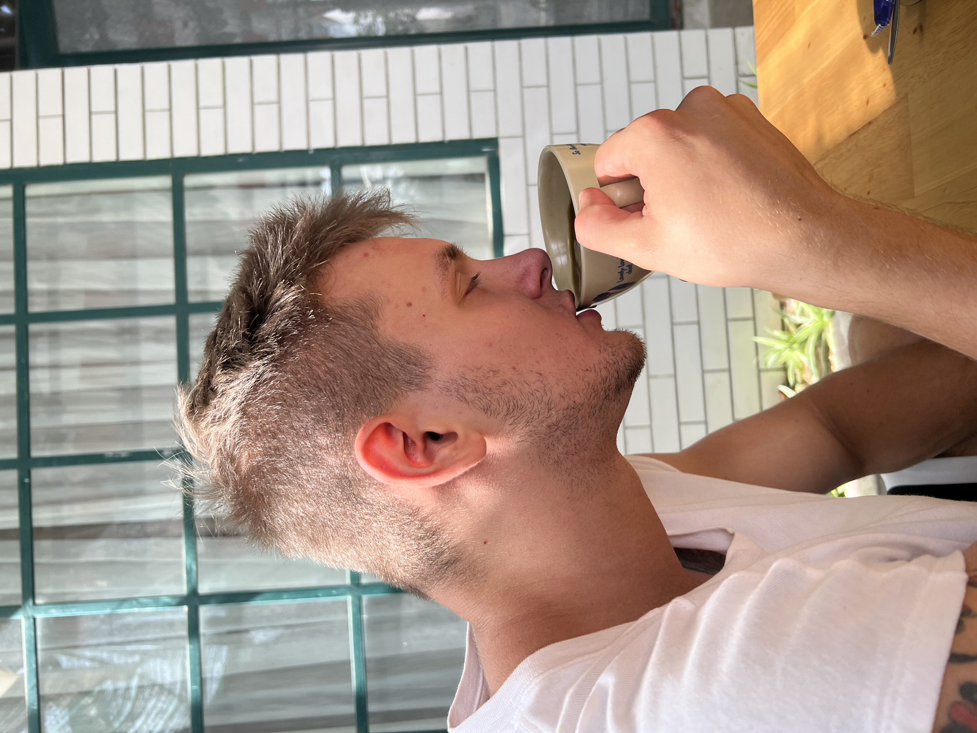 A fair-skinned man is drinking from a beige mug with writing on it. He is wearing a white t-shirt. The background includes a window with a dark green frame and white tile. The setting appears to be indoors, possibly a cafe or restaurant. The location is Bac Nha Trang Ward, Vietnam.