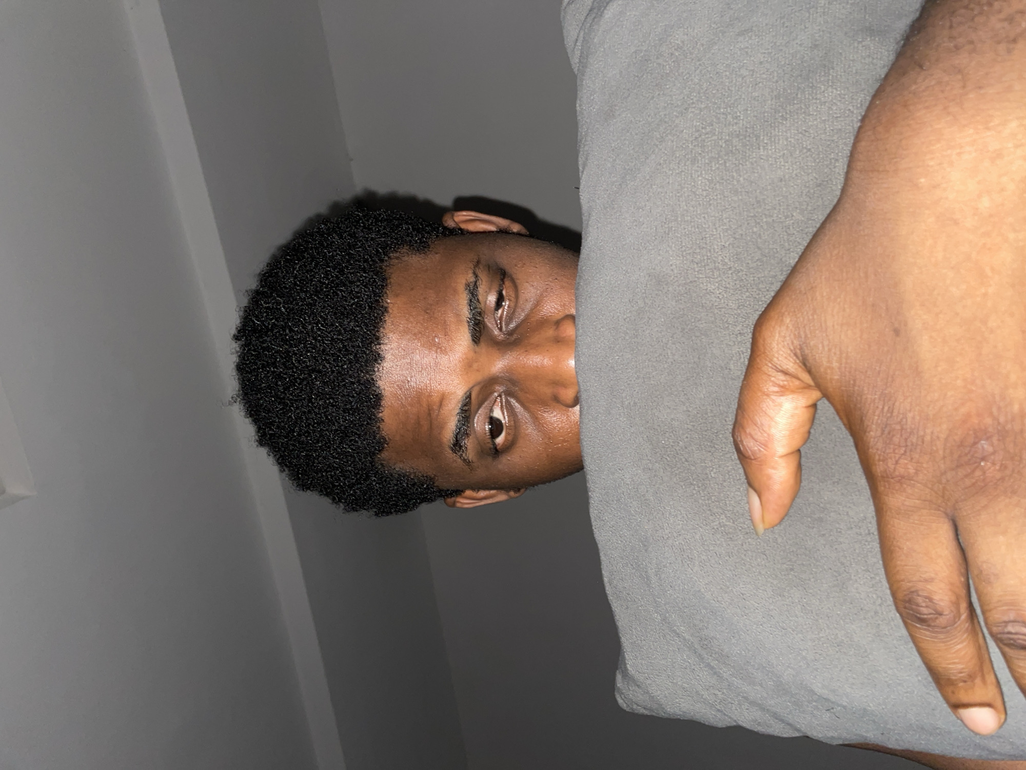 The image presents a rotated close-up shot of a young Black man, peeking from behind a light grey, soft-textured fabric, likely a pillow or blanket. His right eye, brow, forehead, and a portion of his nose and cheek are visible, characterized by smooth, dark skin and short, dark, curly hair. His gaze is directed straight towards the camera, appearing neutral and direct. To the right of the grey fabric, his left hand is visible, resting on the fabric. The background is a plain light grey wall with subtle shadow lines, indicating an indoor setting. The lighting is dim, suggesting the scene takes place in the evening or at night. There is no visible text in the image. The scene captures an intimate moment in an indoor environment in Akwanga, Nigeria.