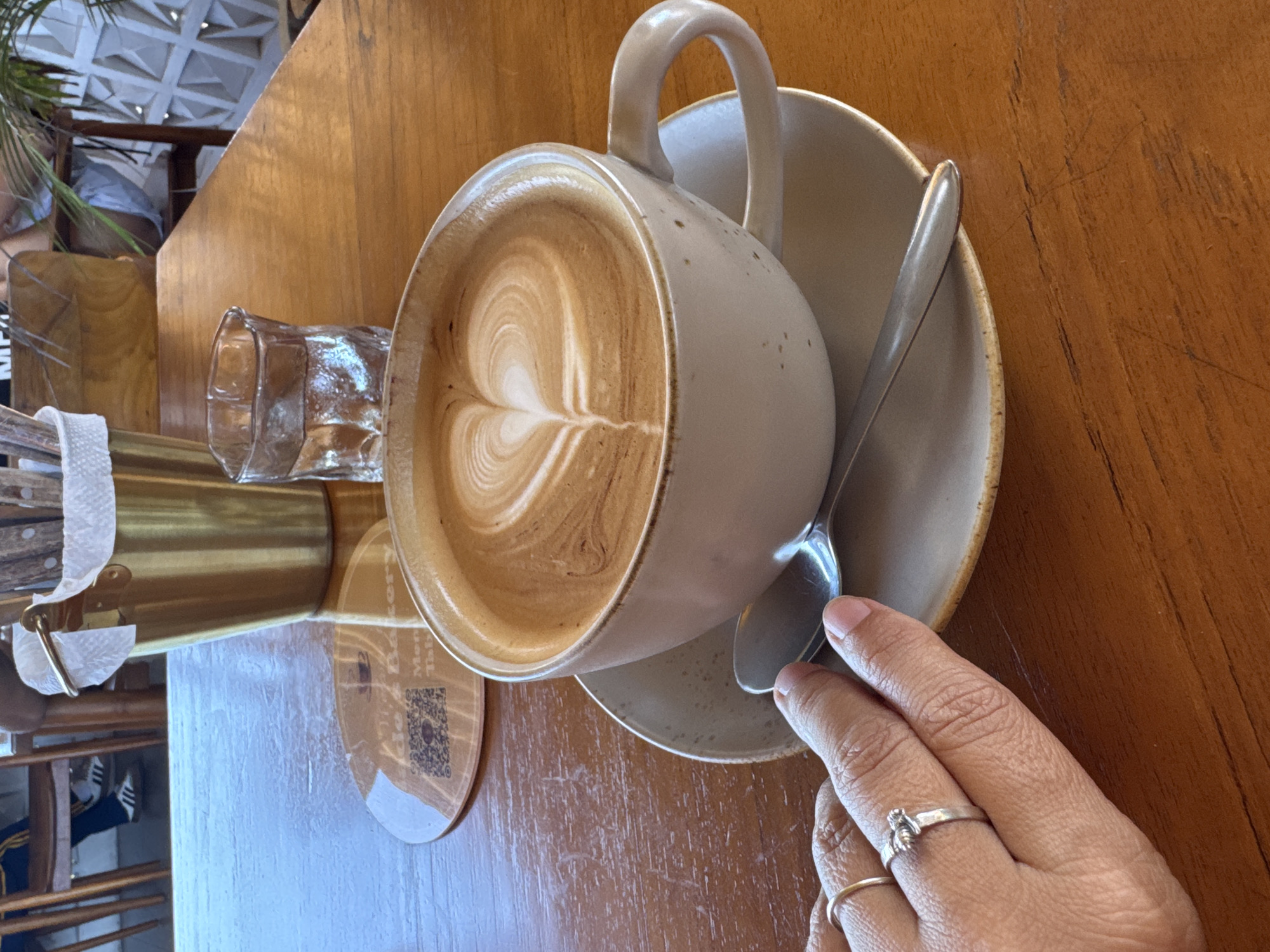 A close-up, slightly angled, and high-resolution image shows a cup of coffee with latte art in the shape of a heart on a wooden table. The cup is made of ceramic, with a slightly speckled texture, and sits on a saucer of similar material. A silver spoon rests on the saucer, near the rim. A hand with two rings (one band, one with a decorative knot) touches the spoon handle. To the left, a small glass of water and a container with cutlery are visible. A coaster with a QR code is under the glass. The wooden table has visible grain. The location is likely a cafe in Kuta Selatan, Indonesia. A person wearing a white top is partially visible in the background.