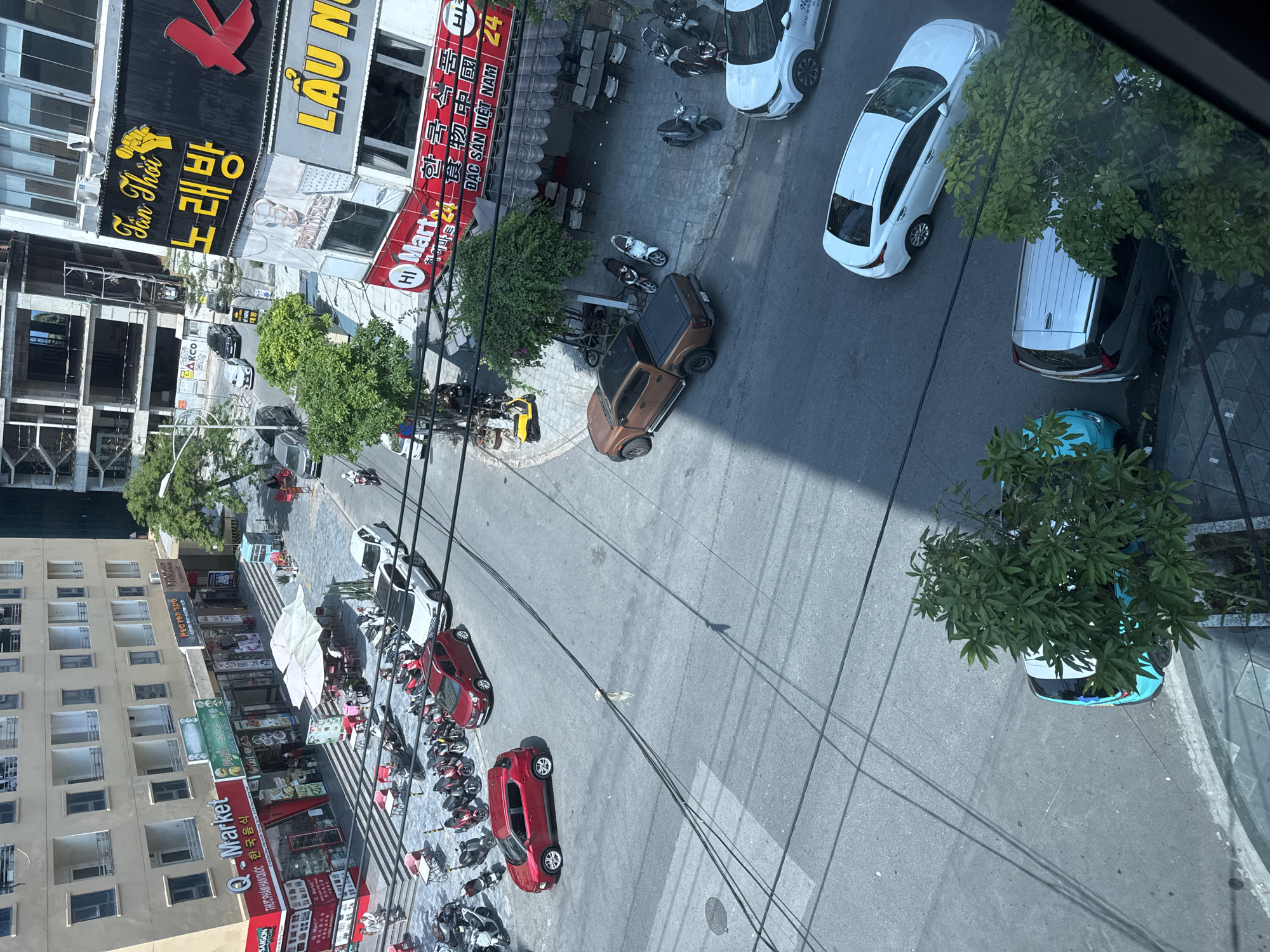 Captured from a high-angle perspective, the photograph presents a street scene in Đà Nẵng, Vietnam. The view features buildings, vehicles, and pedestrians on a city street.
A cluster of buildings occupies the left side of the frame, including a beige-colored structure with several windows and a lower building with a "Q-Market" sign in both English and Korean. Above "Q-Market" are other commercial establishments with signs in Vietnamese and English.
The street is occupied by a variety of vehicles, including cars, trucks, and motorbikes. Several white cars are parked alongside the road. A red car is located on the far left of the street, with motorbikes clustered nearby. A brown pickup truck is positioned perpendicular to the street.
Along the sidewalk, pedestrians can be seen walking and interacting. Outdoor seating areas with chairs and tables are visible near the storefronts. Power lines crisscross above the street.
A building under construction is visible in the background, to the left of the building with the karaoke sign.