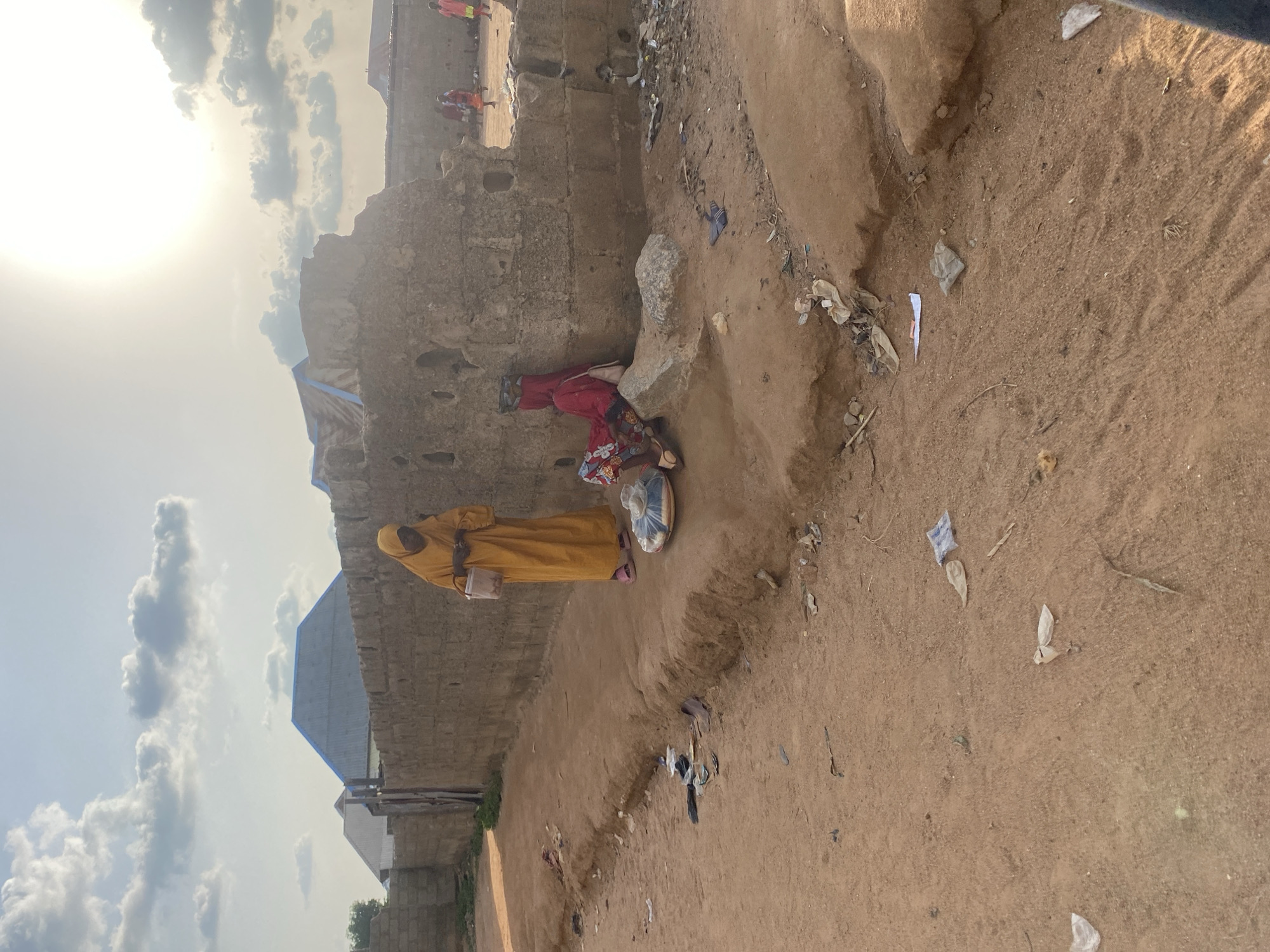 The image depicts an outdoor scene with a sandy ground and a rough, stone wall. The sky is bright with scattered clouds, suggesting daytime. The sun is visible on the left side of the frame, indicating it's likely morning or afternoon.
In the foreground, a person wearing a yellow robe and head covering stands on the sand. They have a light-colored bag hanging from their shoulder. Next to them, a round, flat object, possibly a basket or tray, is placed on the ground. A second person, partially obscured by the wall, is dressed in red and appears to be sitting or leaning against the wall. Further in the background, other individuals are visible near buildings constructed with stone and corrugated metal roofs. The area seems somewhat untidy, with scattered debris and plastic waste on the ground.
The overall impression is of a rural or informal settlement, possibly in a warm climate, given the clothing and the sandy terrain. The presence of multiple people suggests a community setting. The lighting indicates it's a sunny day. There is no visible text in the image.