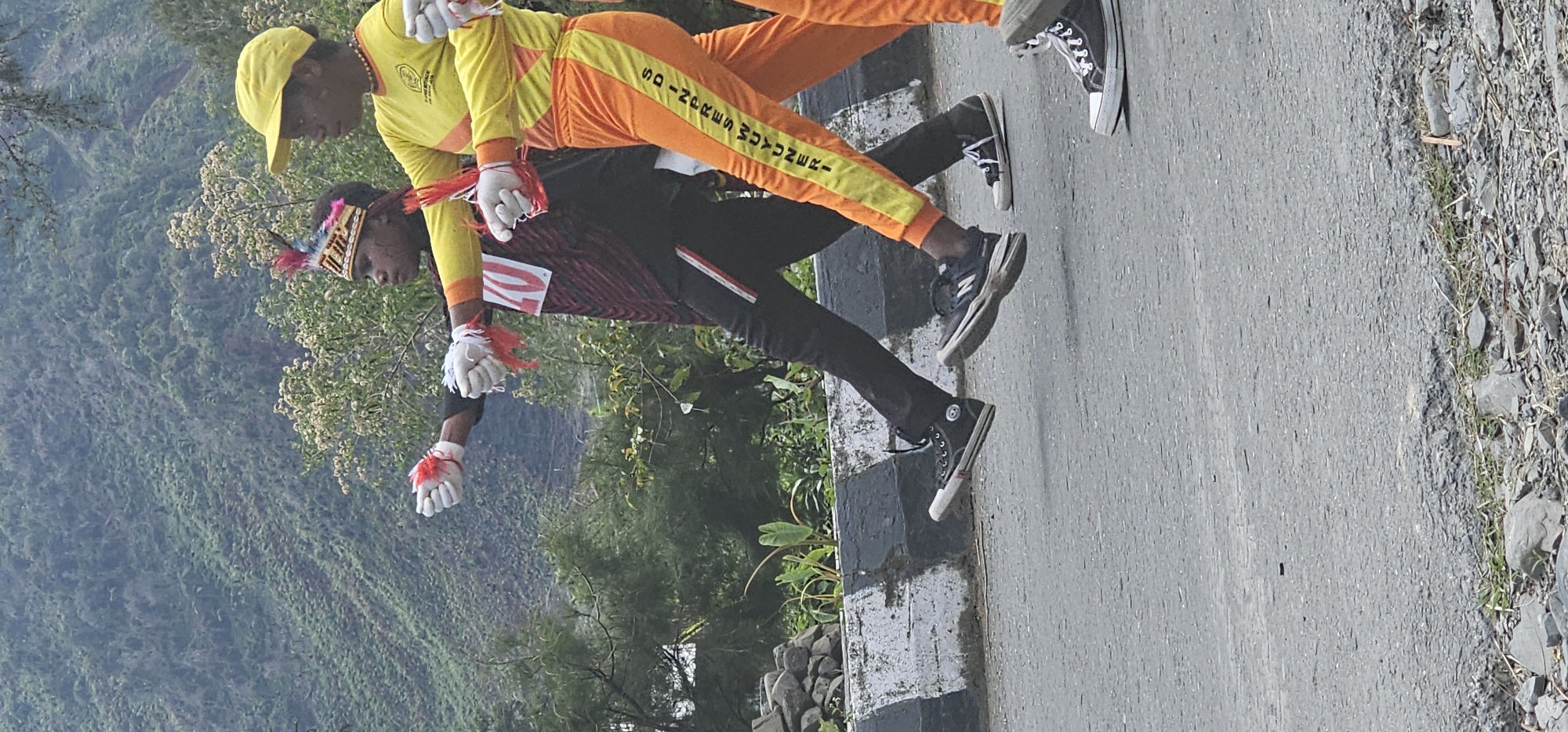 In Puncak Jaya, Indonesia, two individuals are participating in what appears to be a ceremonial march on a paved road. The person in the foreground, on the left, is wearing a dark shirt with red diagonal stripes and the number "40" in red on the front. They have a traditional headdress adorned with colorful feathers. Their hands are gloved in white, with red tassels attached to the wrists. They are wearing black pants with a red and white stripe down the side and black Converse sneakers. The second individual, to the right, is wearing a bright yellow long-sleeved shirt and orange pants with a yellow stripe. They have a yellow baseball cap on. Their hands are also gloved in white with red tassels. The background features a lush, green, mountainous landscape with scattered trees and vegetation. A stone wall with black and white painted sections runs along the side of the road.