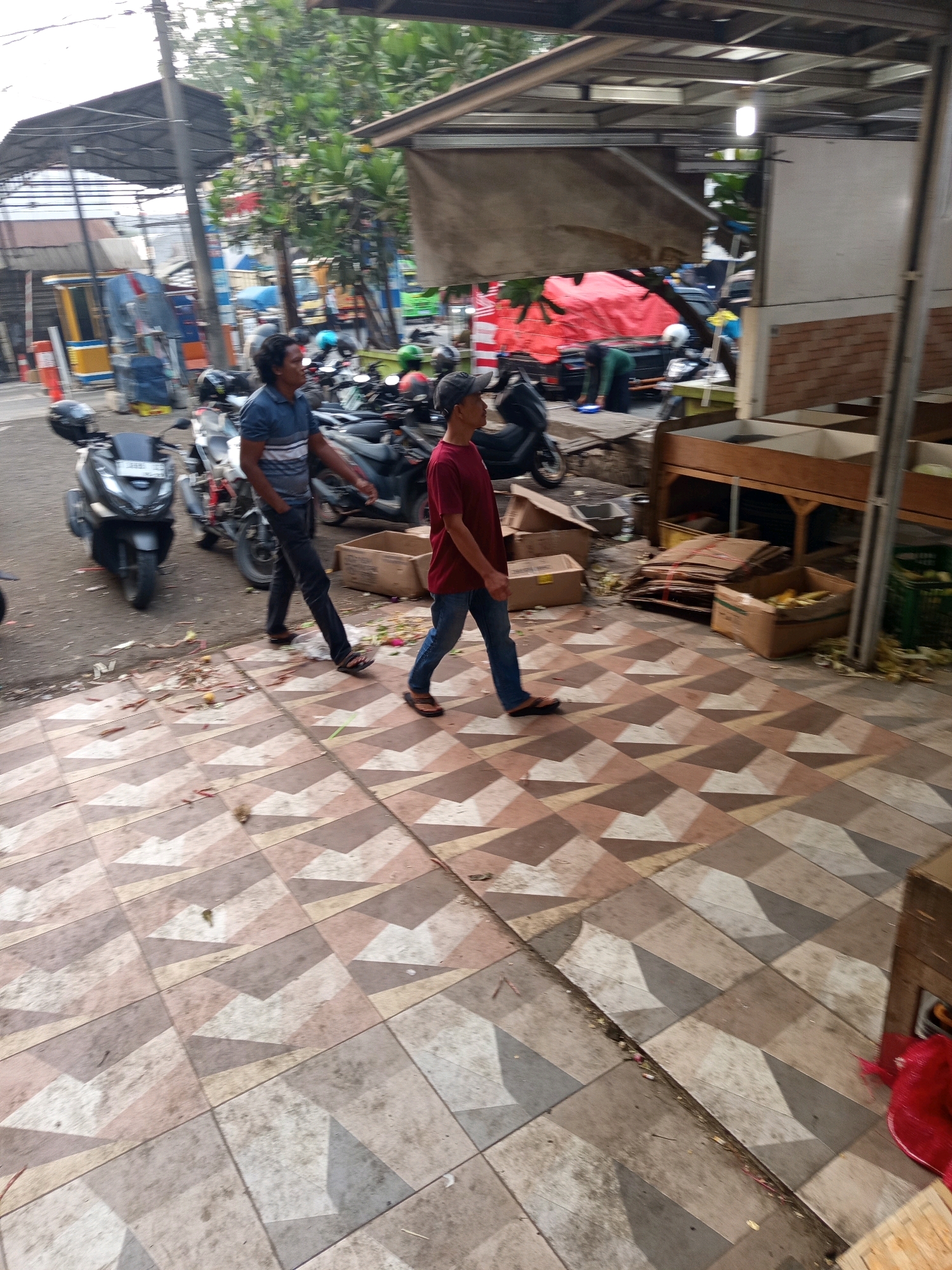 The image depicts an outdoor scene, possibly a market or a roadside stall, in Karawang, Indonesia. In the foreground, two men are walking on a patterned tiled walkway. The man on the left wears a blue and white striped polo shirt and dark pants, while the man on the right wears a maroon t-shirt and blue jeans. To the left of the walkway, several motorcycles are parked, and a portion of a road is visible with traffic cones. In the background, there are structures with awnings and stalls displaying what appear to be produce, such as corn in a cardboard box. The environment seems to be somewhat cluttered, with discarded cardboard boxes and debris on the ground. Trees and overhead wires are also visible, suggesting an urban or semi-urban setting. The lighting indicates it is daytime.
