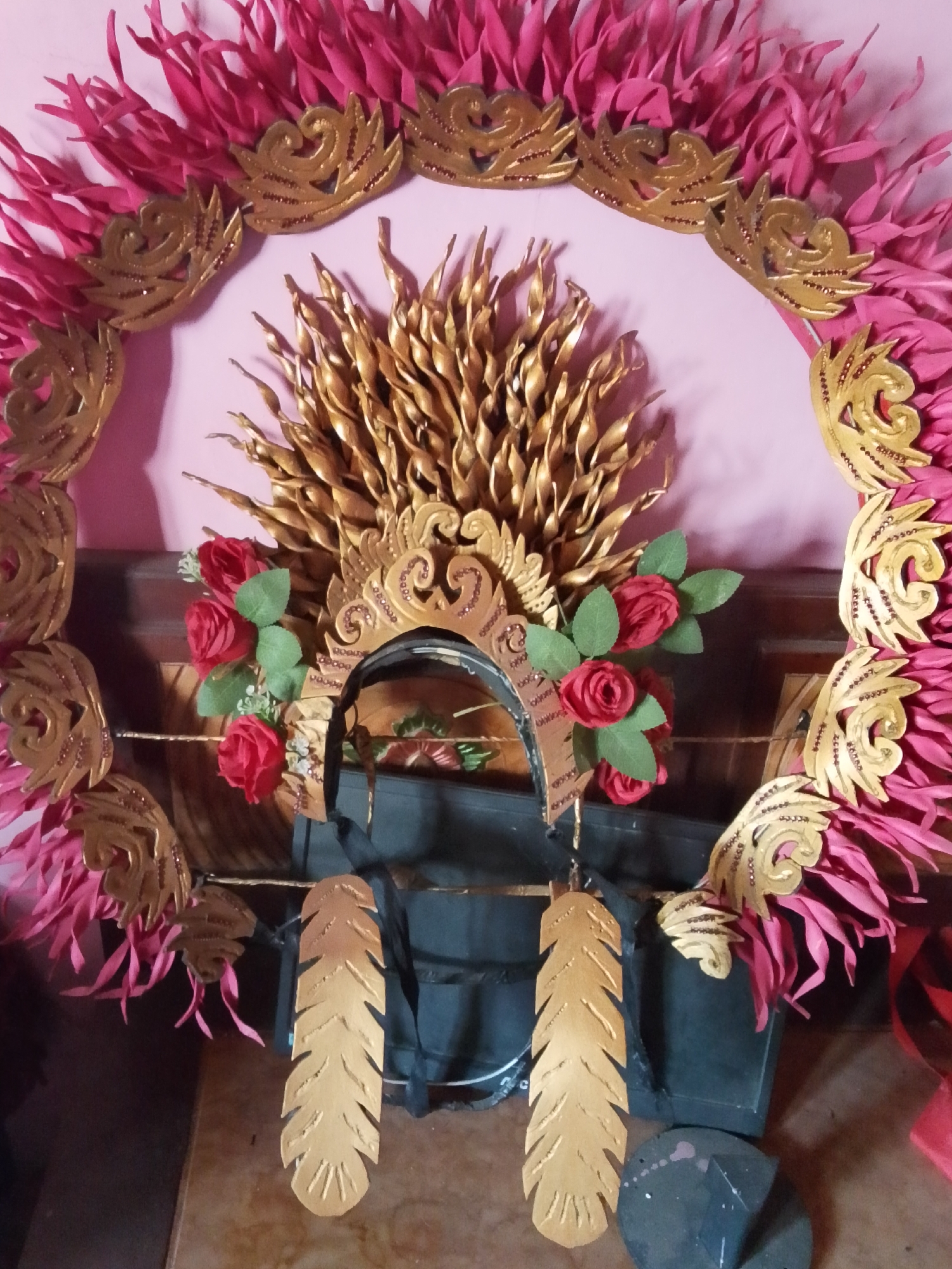 The image shows a detailed view of a traditional Balinese headdress, likely for a dance performance. The headdress is predominantly gold and pink, with intricate gold leaf patterns and vibrant pink feather-like accents forming a circular halo. In the center, a darker, likely black, fabric object is partially visible, with a gold crown-like embellishment and several realistic red roses with green leaves adorning the sides. Two gold feather-shaped ornaments hang from the headdress, one on each side. The headdress is displayed against a light pink background, with a wooden table and furniture visible in the foreground, suggesting it is in a domestic setting. The overall impression is of a handcrafted cultural artifact, possibly a prop for a ceremony or performance in Bulurejo, Indonesia, given the context provided. There are no people visible in the image.