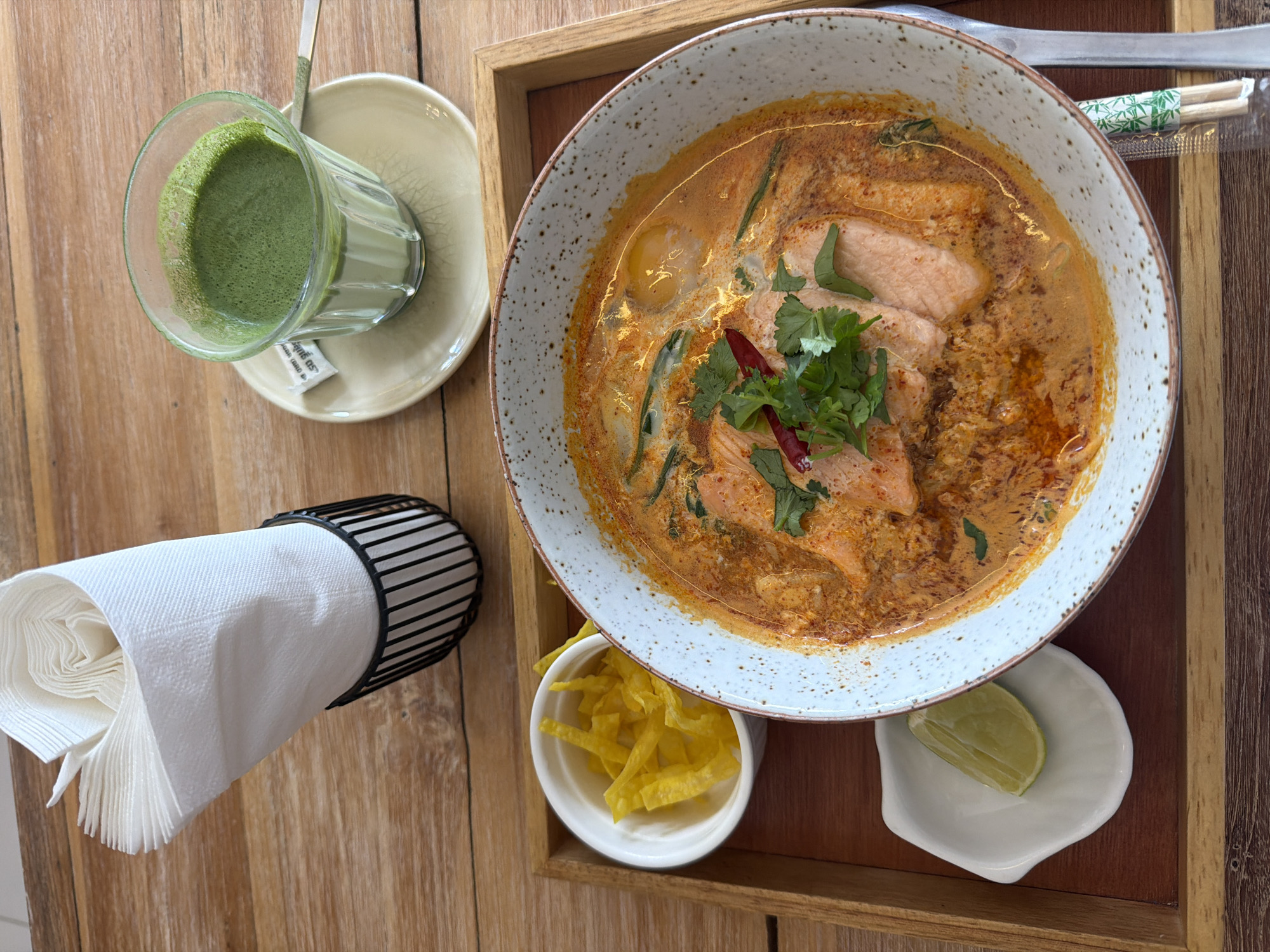 A top-down, high-angle medium shot captures a wooden table surface at a restaurant in Koh Phangan, Thailand. On the table is a wooden tray containing a bowl of steaming salmon curry soup, a small white dish with yellow fried noodles, a small white dish with a lime wedge, and a glass of green matcha tea on a saucer with a sugar packet. To the left of the tray, a black wire napkin holder is filled with white napkins. The scene is brightly lit, suggesting it's daytime.