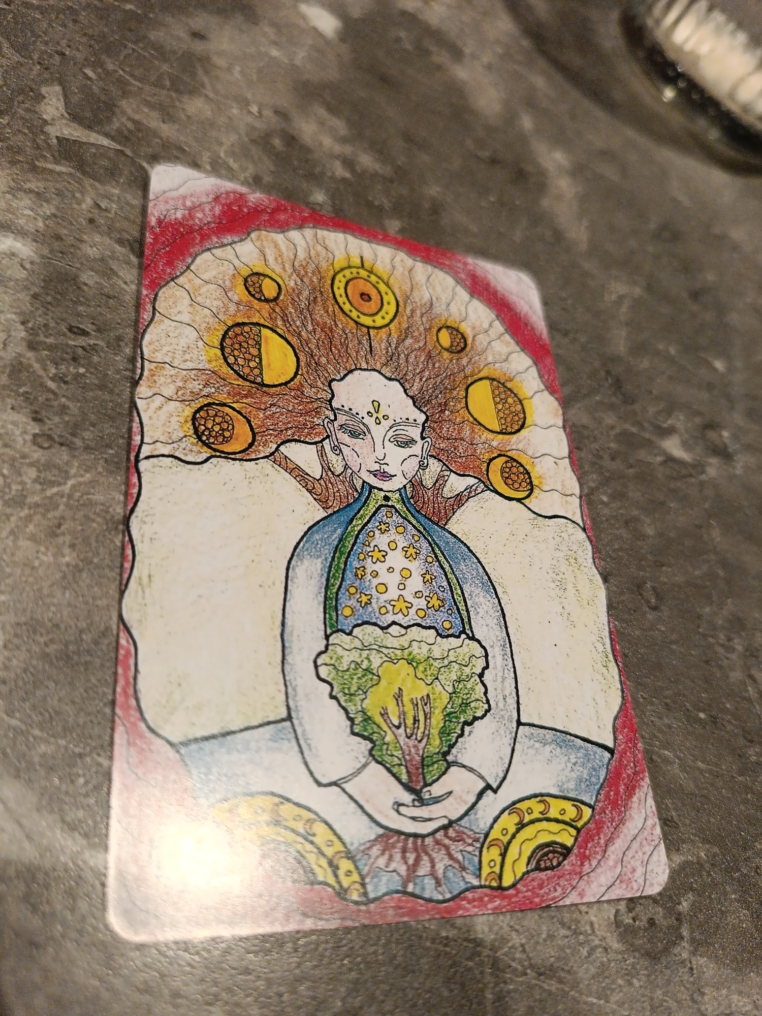 The image is of a tarot card depicting a woman. Her hair fans out behind her head, containing several circular, golden symbols, some of which resemble phases of the moon. She has pale skin, delicate features, and three dots on her forehead. She wears a blue robe with yellow stars, and green trim. She is seated and holds a small, leafless tree or plant with exposed roots in her hands. The background is white with red outlining the perimeter of the card. The card is lying on a grey, textured surface. There is no evidence to suggest it is in Chaikovsky, Russia.